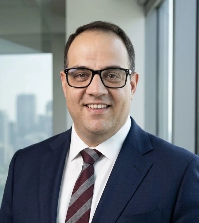 A man in a suit and tie smiling, wearing black glasses, standing indoors near windows with a cityscape in the background.