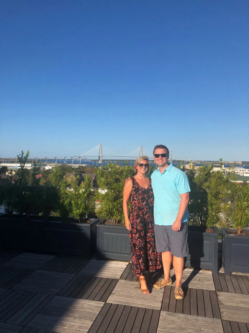 A smiling couple standing on a rooftop terrace with potted plants, a cityscape, and a bridge in the background on a sunny day.