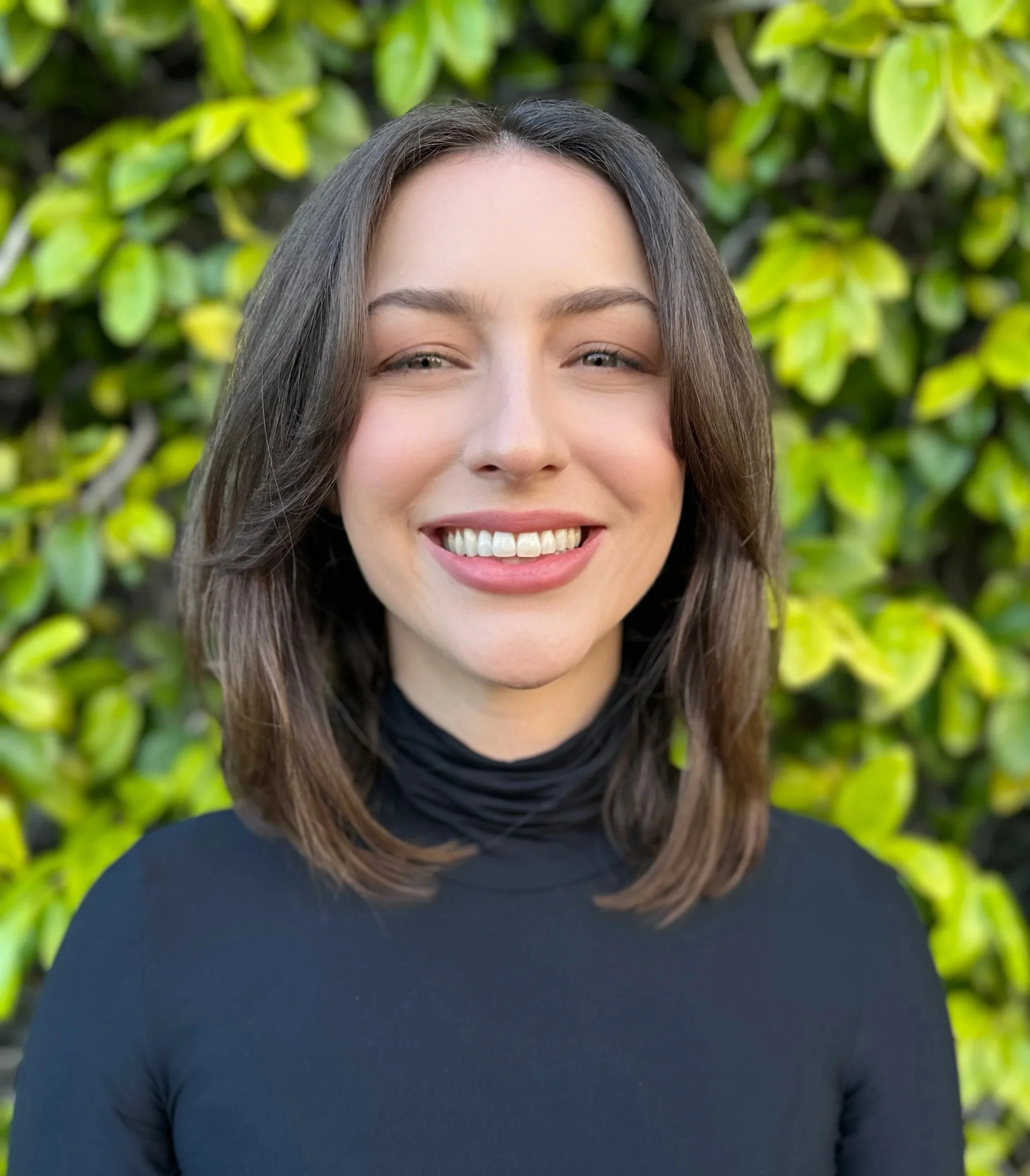 Close-up of a woman smiling, with straight brown hair and light skin, standing outdoors with green leaves in the background, wearing a black turtleneck.