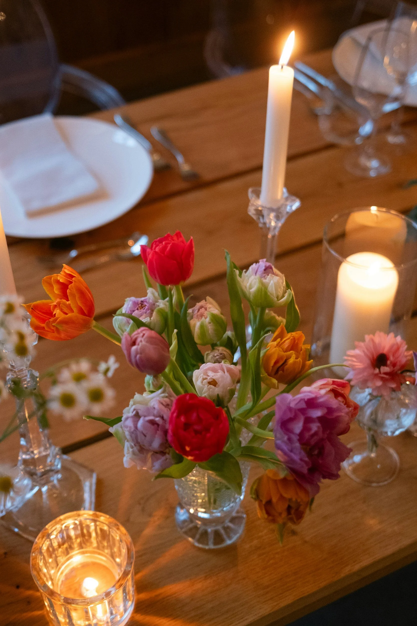 A dining table decorated with a colorful assortment of tulips and other flowers in glass vases, lit candles in various holders, and tableware including plates and cutlery.