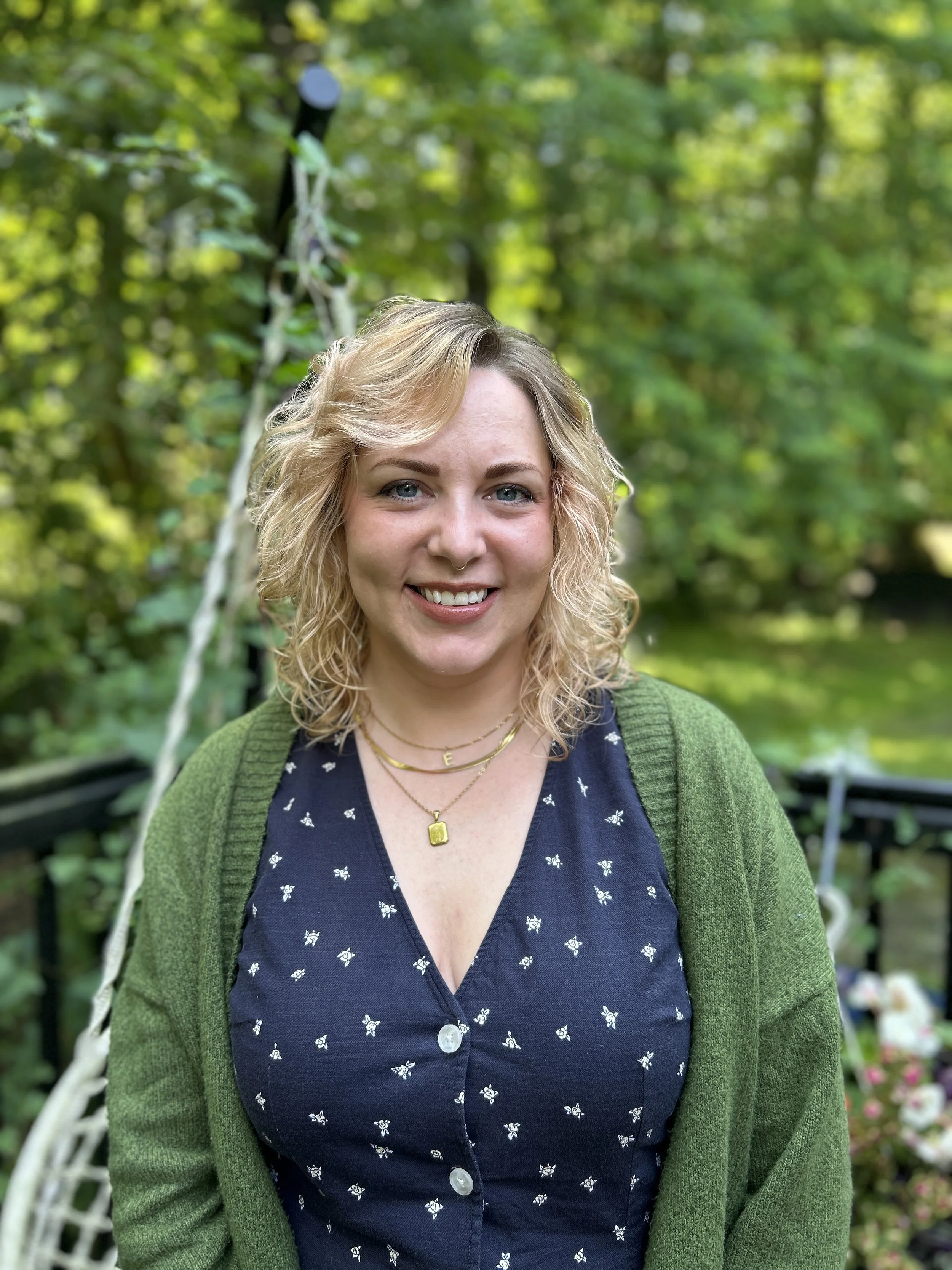 A woman with curly blonde hair smiling outdoors in a garden, wearing a dark blue dress with a butterfly pattern, layered with a green cardigan, and gold jewelry.