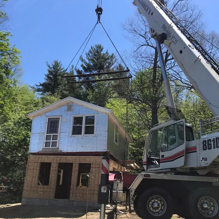A crane lifting a small house under construction, with a blue sky and trees in the background.
