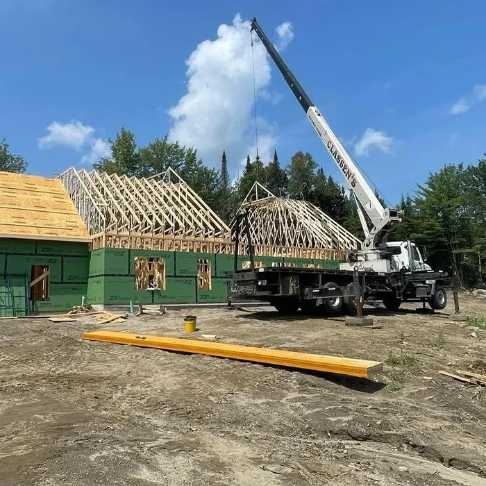 A construction site with a house in the framing stage, showing wooden roof trusses, and a crane truck on site.