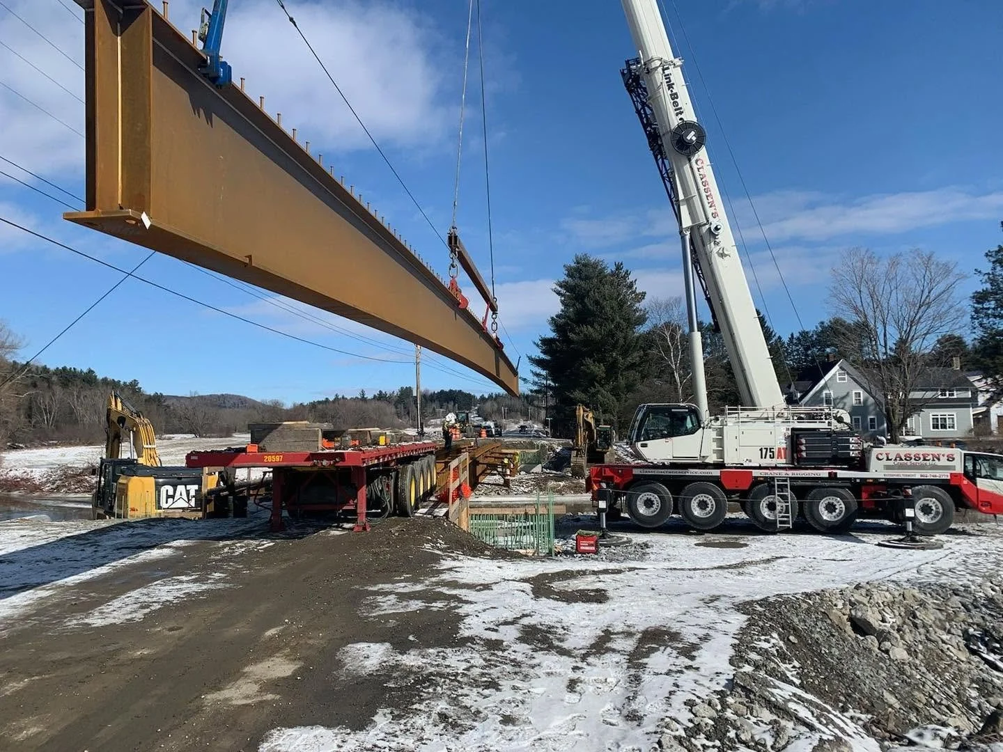 A large crane lifting a steel beam during a construction project outdoors on a partly snowy day with houses and trees in the background.