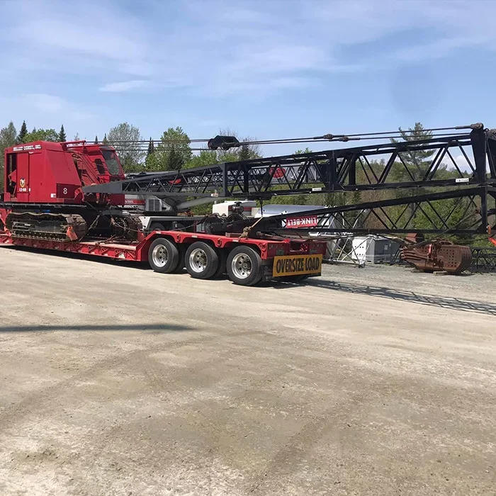 A large crane with a red base and black arm on a truck bed, marked with an 'Oversize Load' sign, parked on a gravel lot with trees and blue sky in the background.