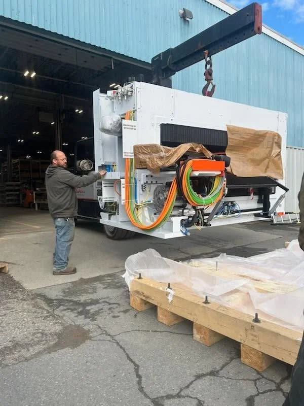 A man operating a control panel next to a large machine wrapped in paper outside a warehouse. The machine has multiple colored cables and a crane nearby.
