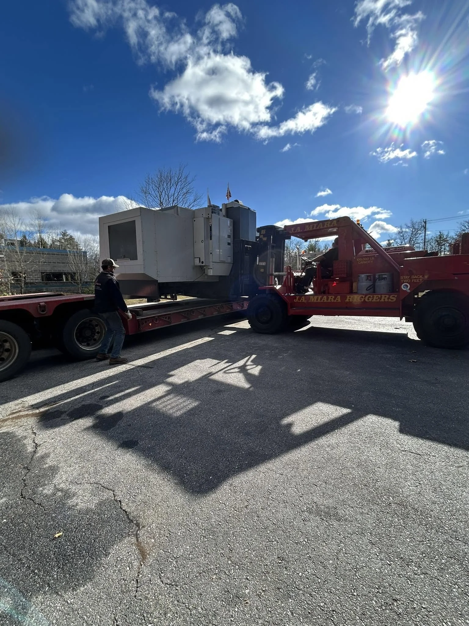 A flatbed tow truck carrying a large white machine under a clear blue sky with scattered clouds and the sun shining brightly.