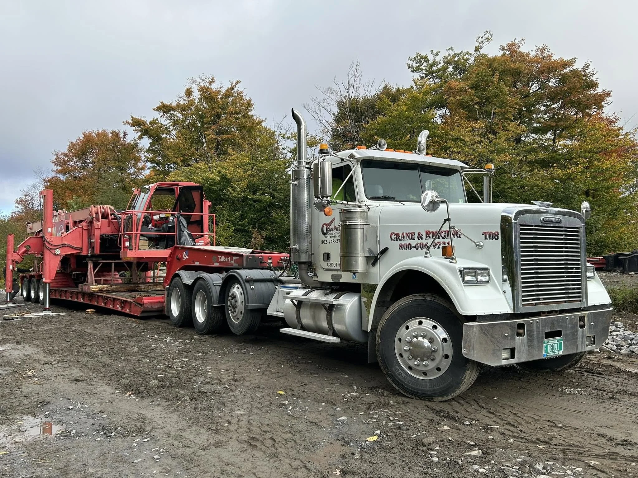 A large white truck with a red construction attachment parked on a muddy surface, with trees showing fall foliage in the background.