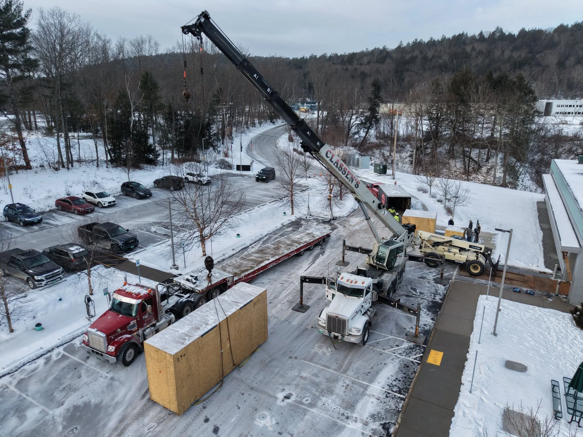 A construction crane lifting large wooden panels in a snowy parking lot with cars and trees in the background.