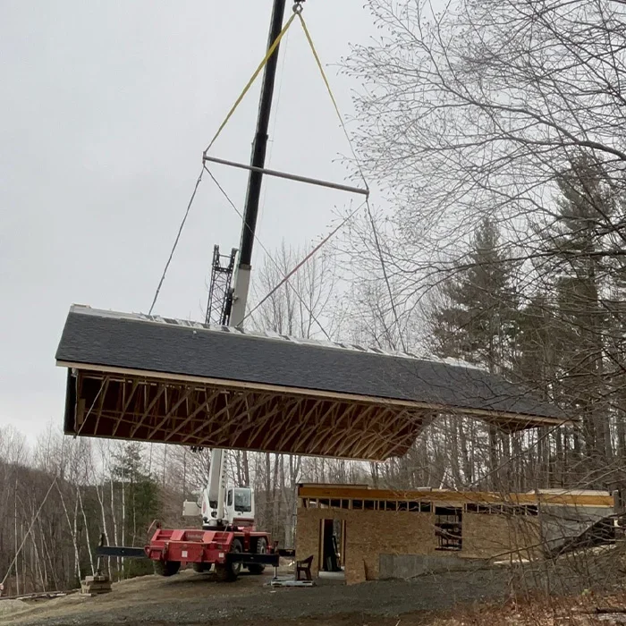 A house addition with a roof being lifted by a crane during construction, surrounded by trees in a wooded area.
