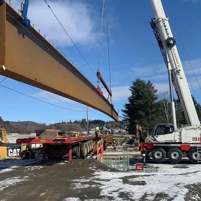 A construction crane lifting a large steel beam on a construction site with snow and trees in the background.