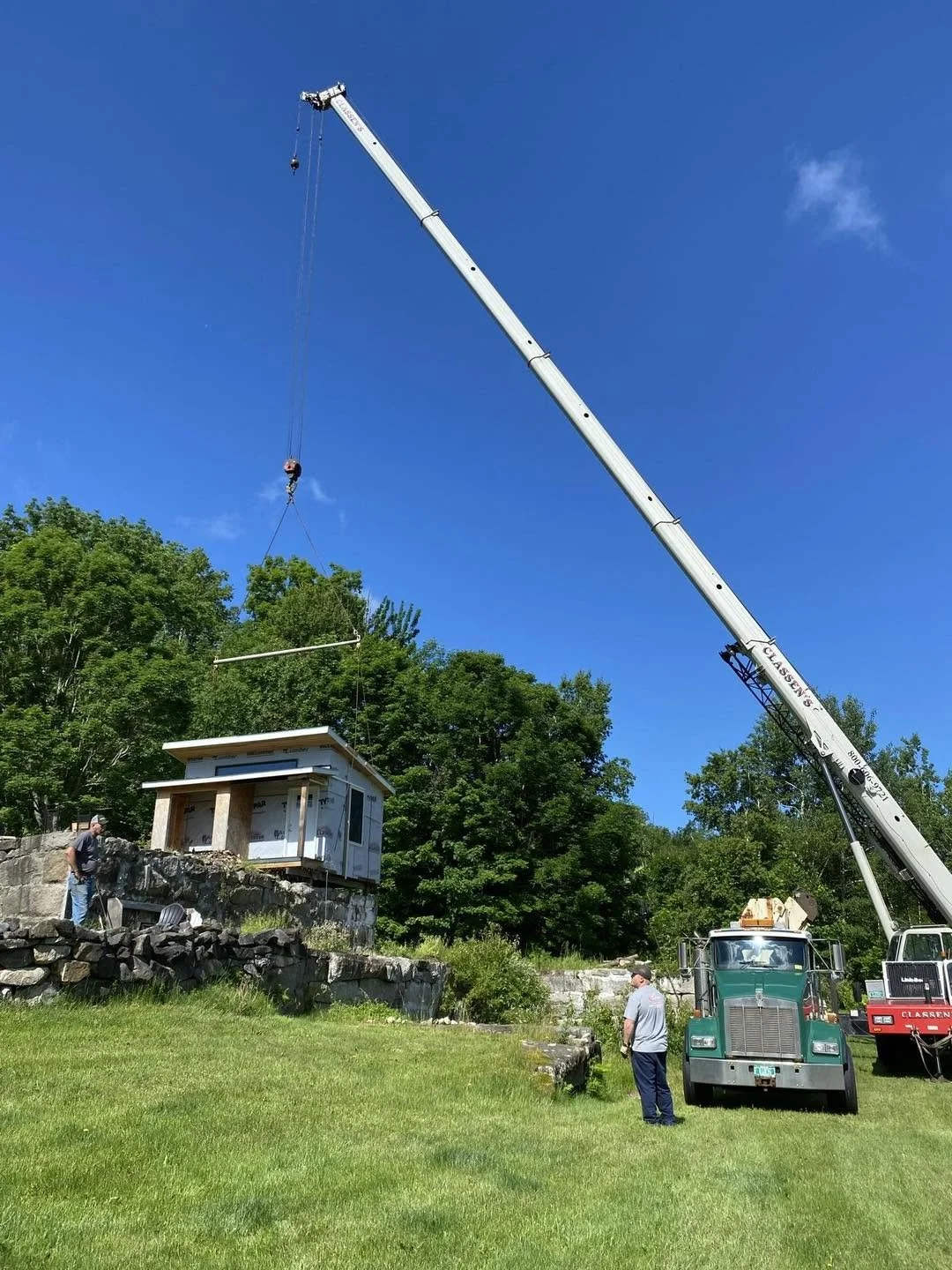 A crane lifting a small house or shed onto a stone foundation with two people standing nearby and green trees in the background on a sunny day.