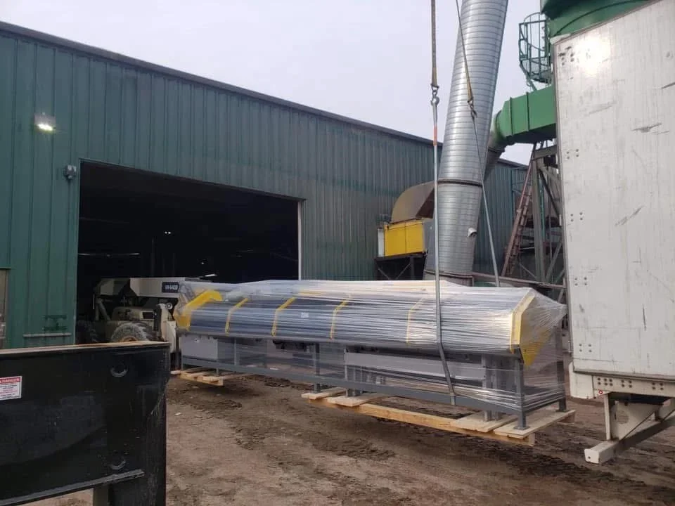 Industrial outdoor scene with a green warehouse, large ductwork, and a pallet carrying a long metal machine wrapped in plastic.