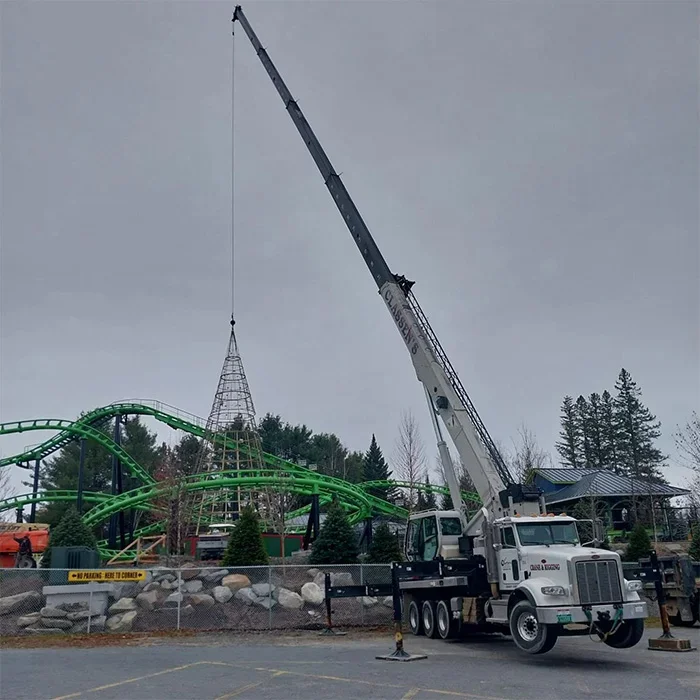 A large crane lifting a Christmas tree decoration at an amusement park with a roller coaster in the background.