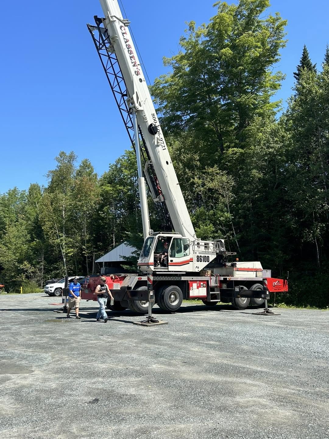 A large white crane with red accents and black supports is parked on a gravel lot, with green trees and blue sky in the background. Three people are standing near the crane, two wearing baseball caps and casual clothing, the third wearing a blue shirt, khaki shorts, and a backpack.