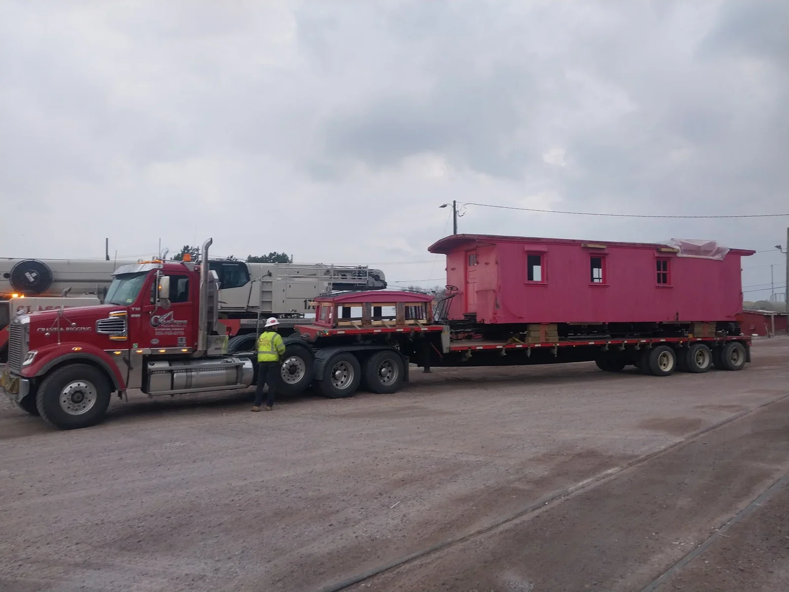 A large red truck is transporting a pink house on a flatbed trailer, with a worker in a yellow safety vest and a white hard hat standing nearby. The scene appears to be outdoors on a cloudy day.