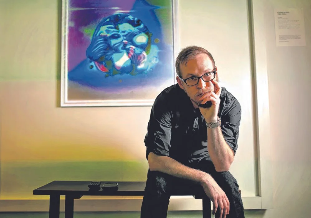 Kyle Van Osdol, with glasses, sits on a stool in front of his abstract artwork located in the Underground tunnels in Oklahoma City.