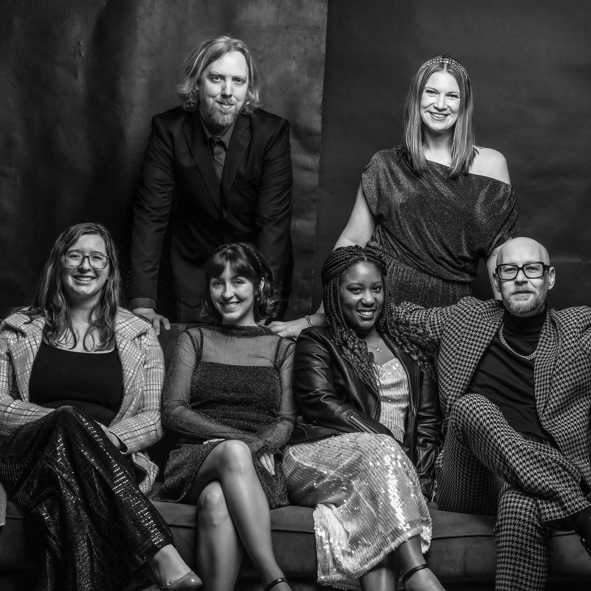 Black and white photo of a diverse group of people sitting and standing together, smiling, in a studio setting. The group includes men and women of different ages and ethnicities, dressed in stylish clothing, with a plain dark background.