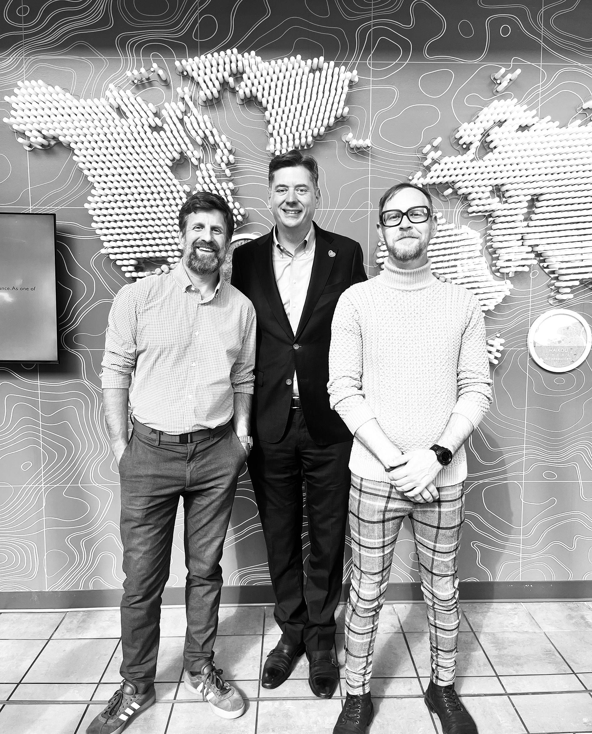 Three men standing in front of a world map wall display at an indoor location, smiling for the photo.