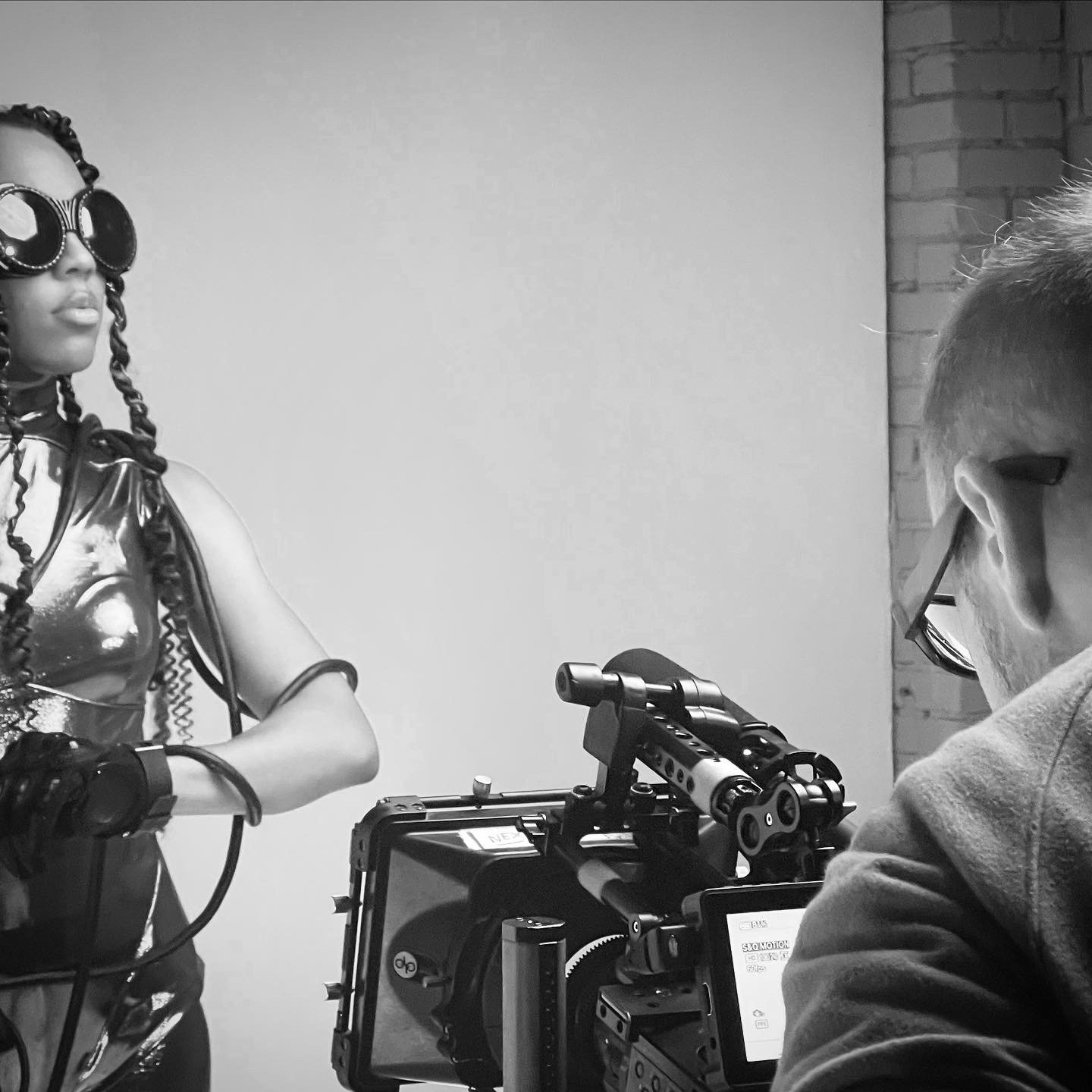 Behind-the-scenes photo of a woman with braided hair and large sunglasses, being filmed by a man with glasses and a camera on a tripod.