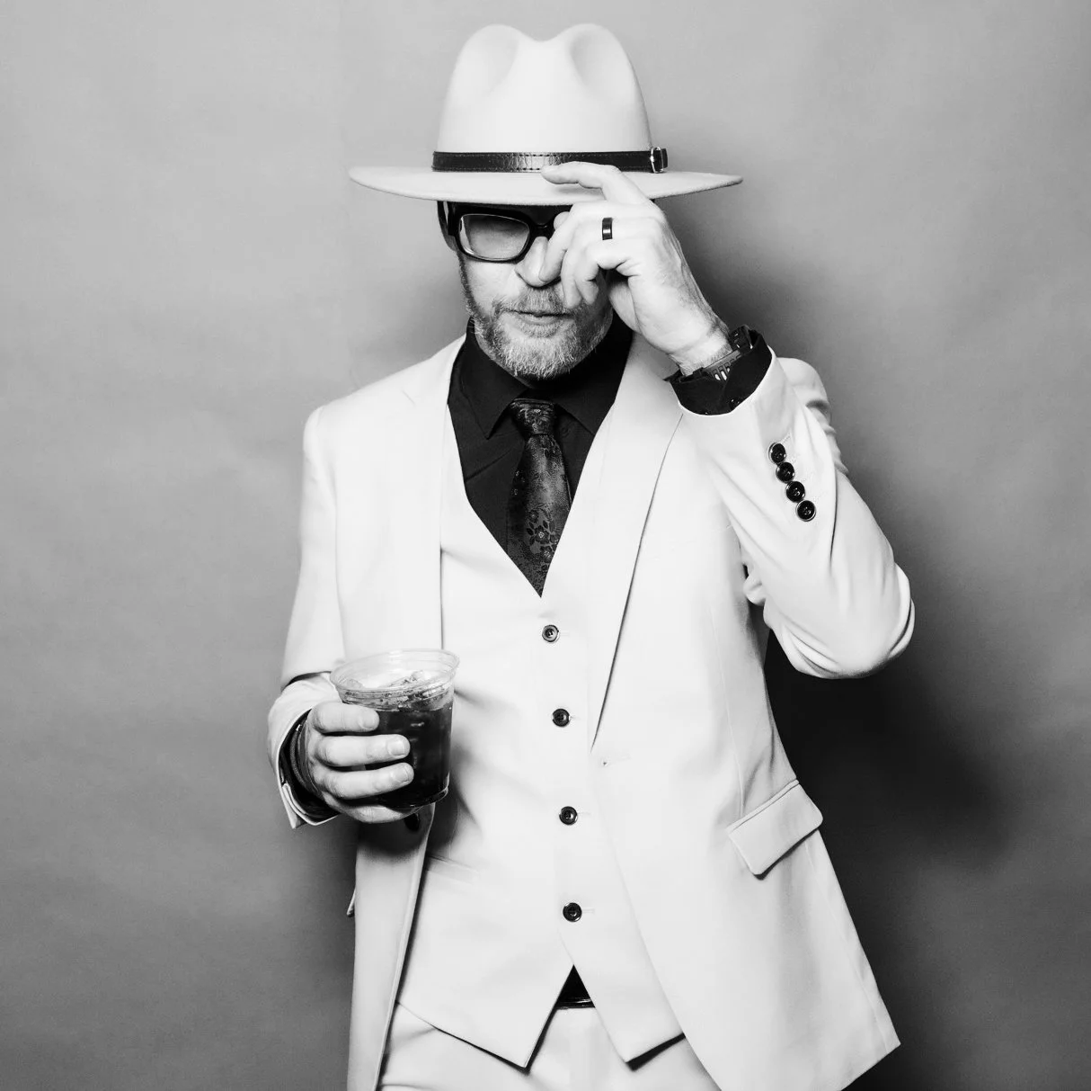 Kyle Van Osdol in an all yellow suit man wearing a wide-brimmed hat, glasses, and a ring on his finger, holding a drink in his left hand, adjusting his hat with his right hand, standing against a plain background at Six Twelve's Mardi Gras Ball.