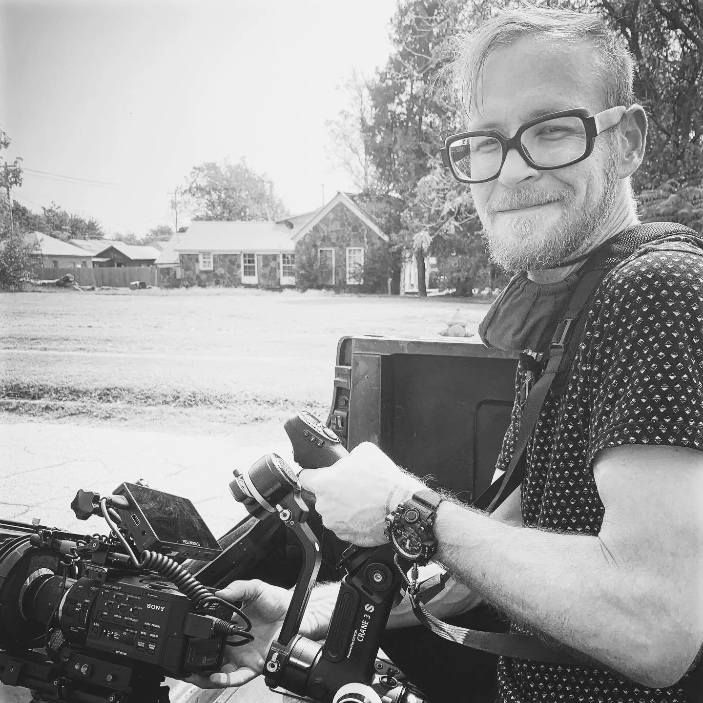 A man with glasses and a beard operating a professional camera mounted on a gimbal, outdoors in front of a residential neighborhood, smiling and looking to the side.