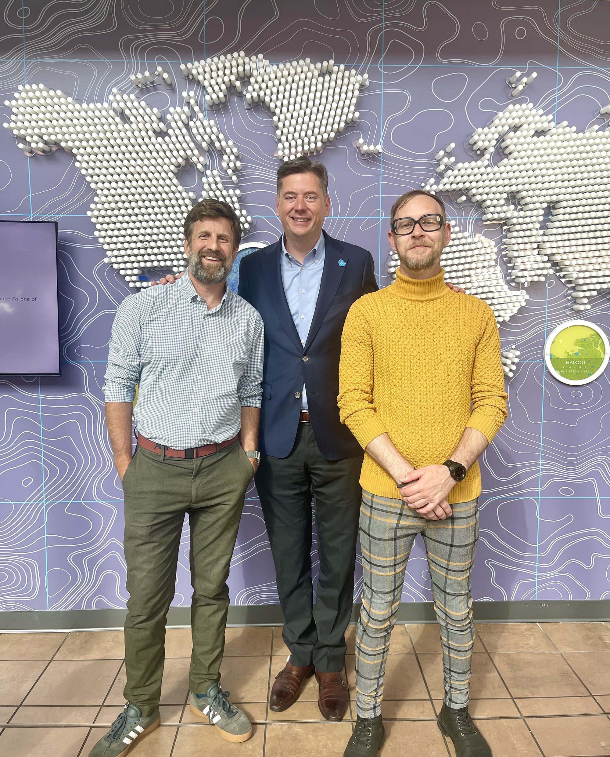 Kyle Van Osdol, Scott Henderson, and Mayor David Holt standing together in front of a world map installation locataed at Will Rogers Airport in Oklahoma City.
