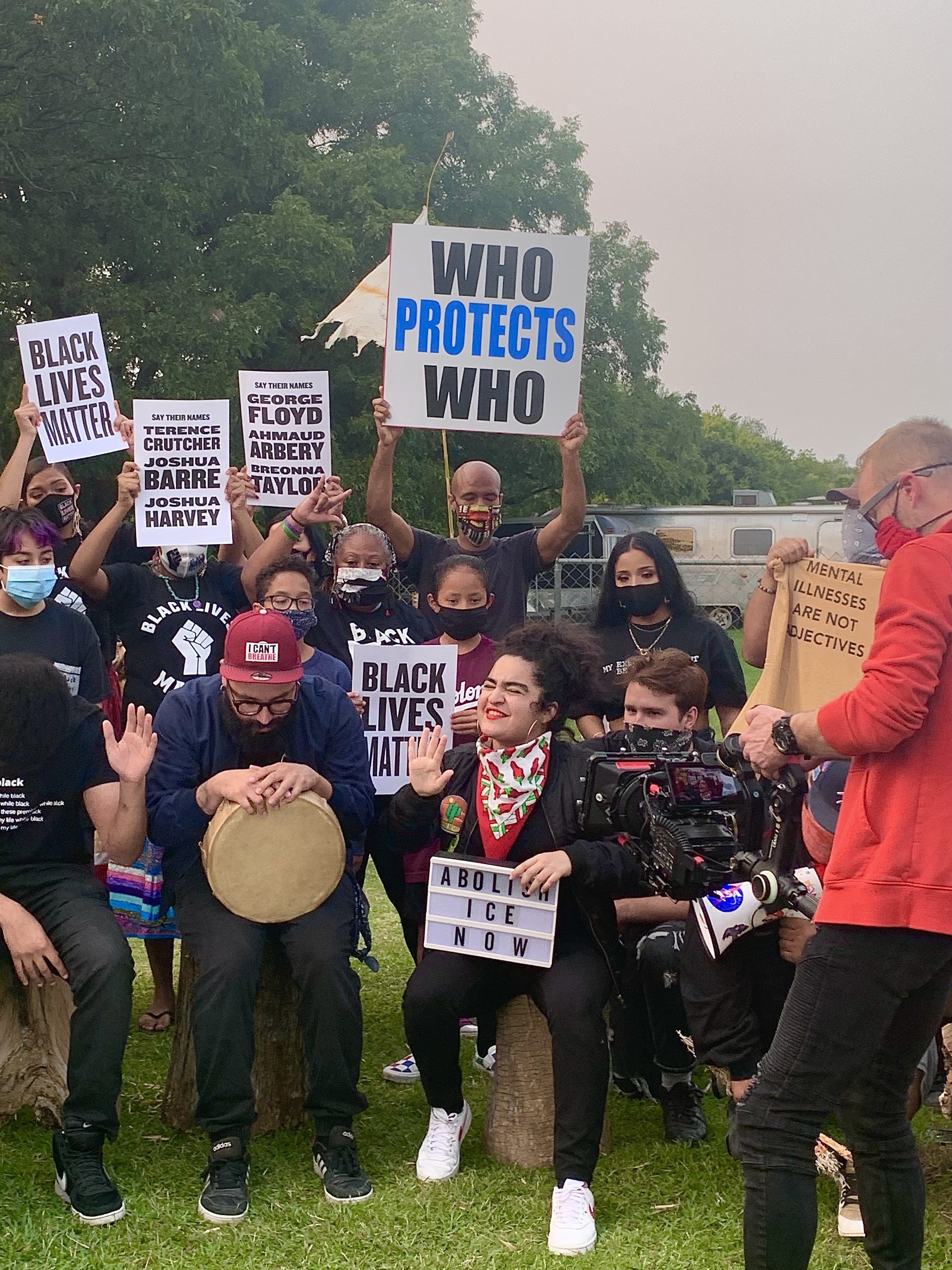 Group of diverse people protesting outdoors, holding signs supporting Black Lives Matter, police accountability, and justice, with trees and a fence in the background.