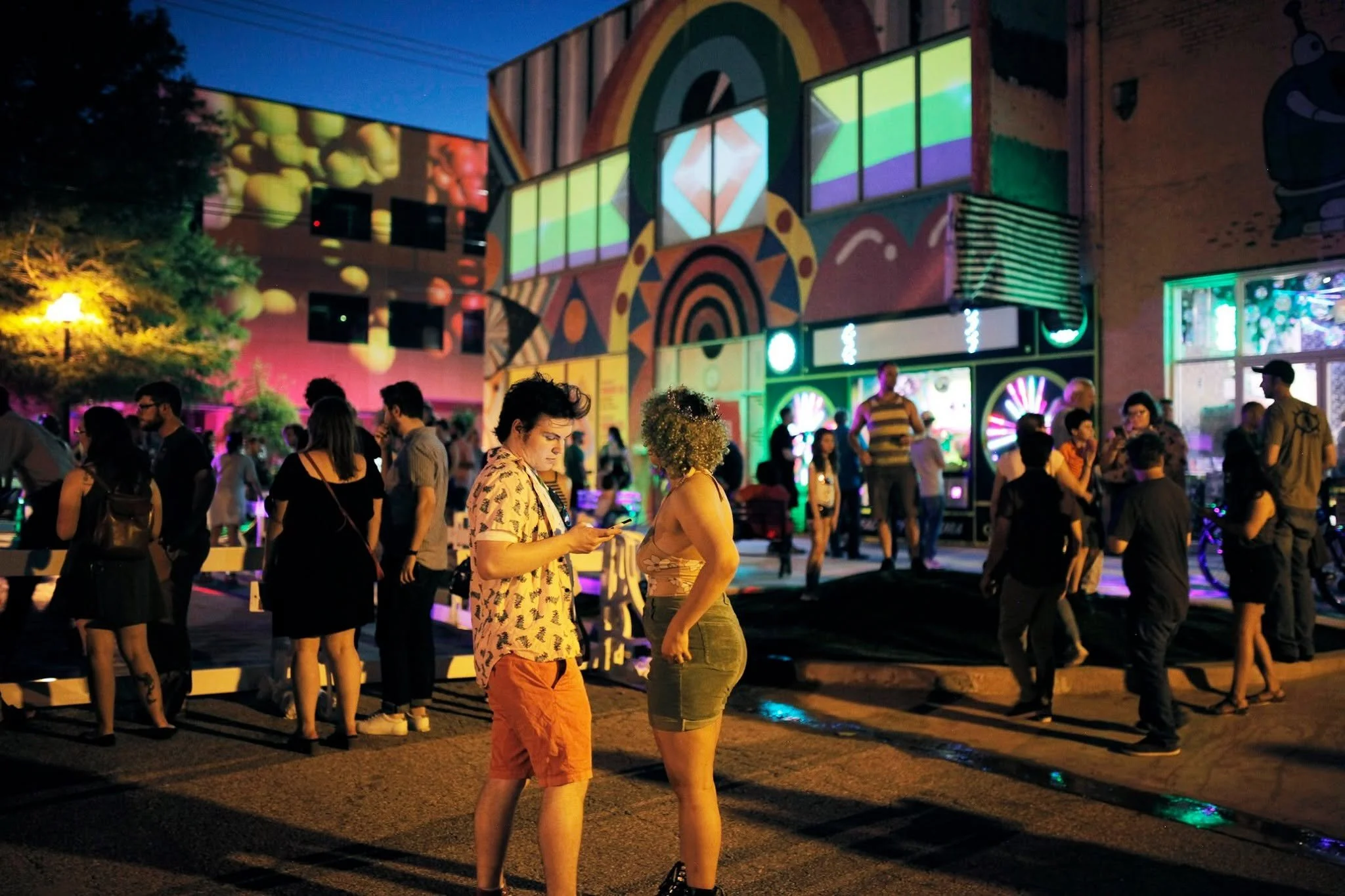People gathering outdoors at night with colorful building projections and neon lights, some talking and others walking, in an urban setting.
