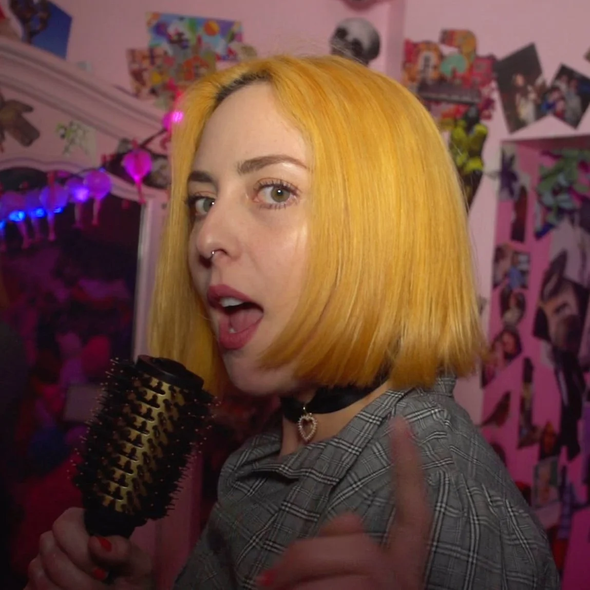 Young woman with bright yellow hair and septum piercing holding a hairbrush, pointing towards the camera in a decorated room with pink walls and photos.