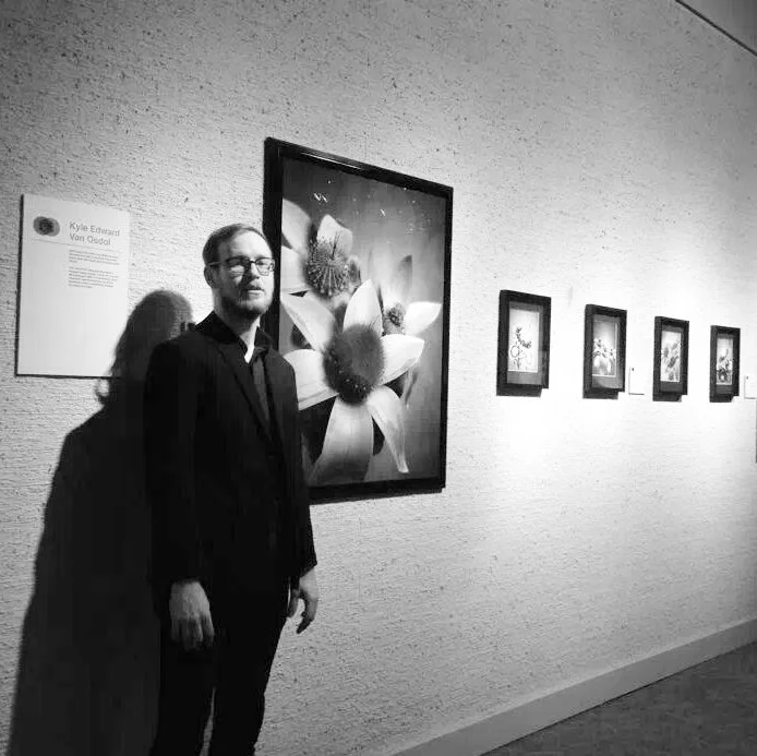 A man in black clothing and glasses standing in an art gallery, with framed photographs or artwork on the wall behind him, including a large floral image.