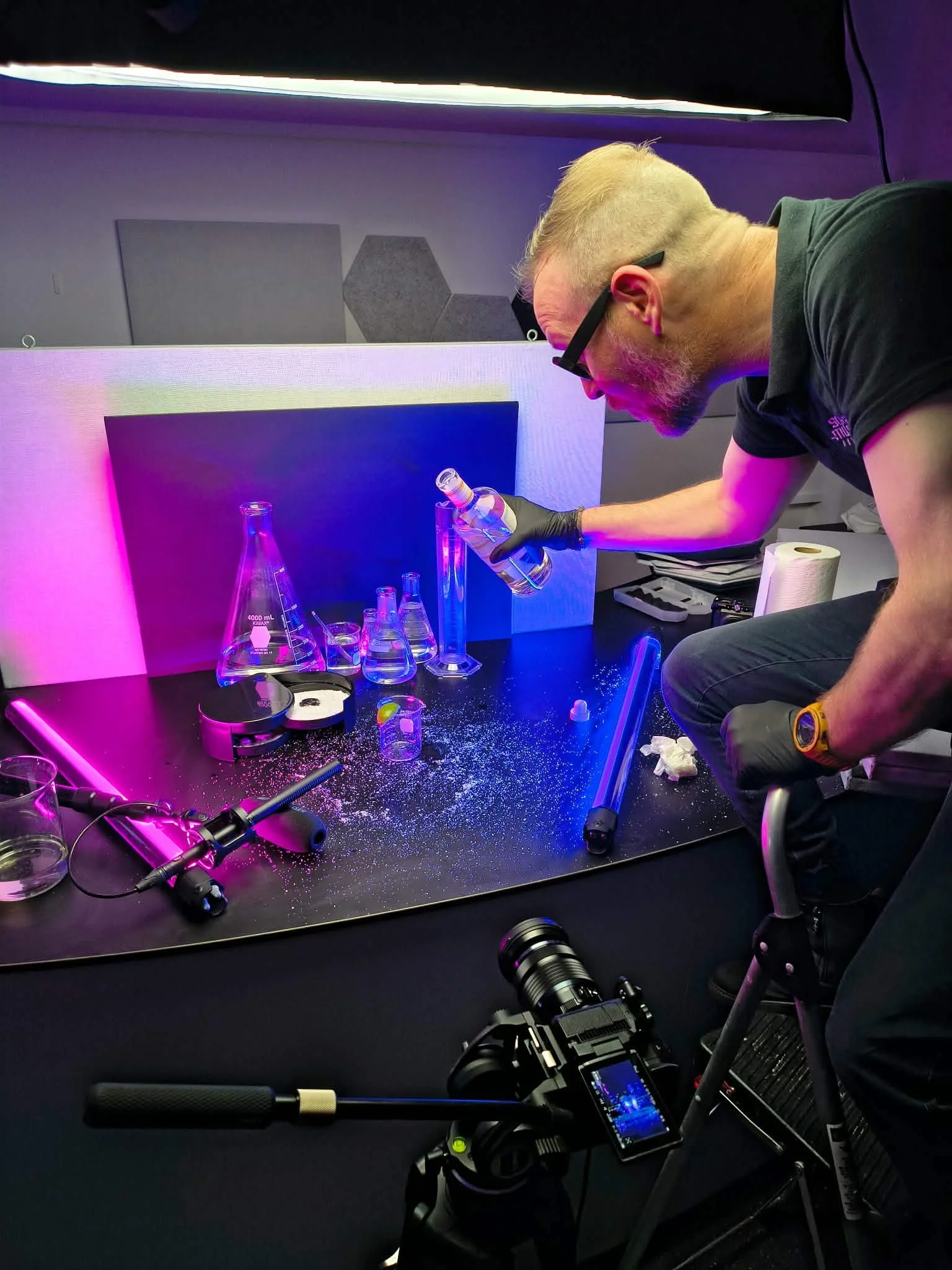 Kyle Van Osdol is pouring a clear liquid from a bottle into a test tube on a set at Science Museum Oklahoma with purple and pink lighting. A camera on a tripod is positioned near the man, and sound or recording equipment is nearby.