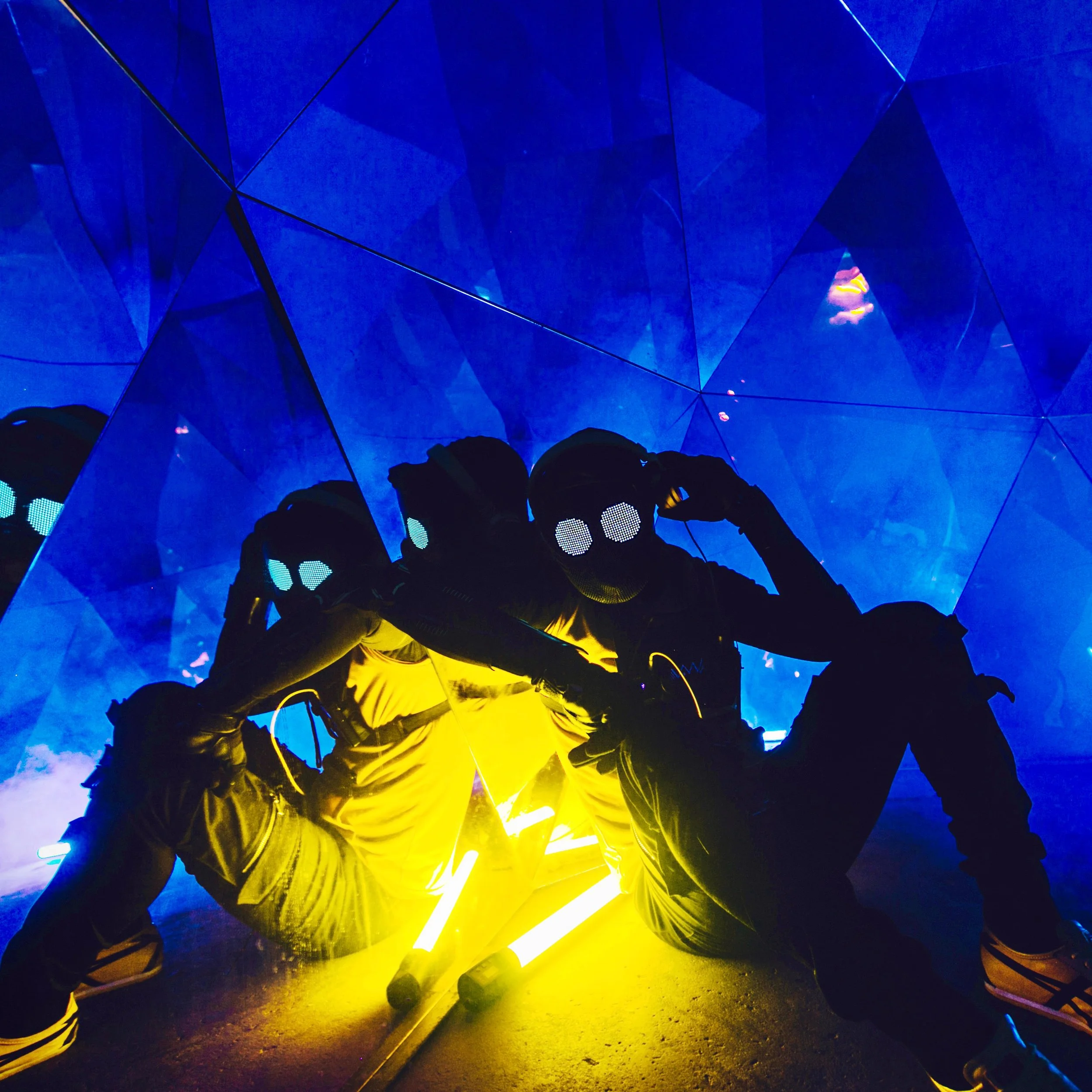 Kyle Van Osdol is seated on the ground and wearing a mask, dressed up as his musical moniker, Dr. Van, sitting in an illuminated sculpture with bright yellow lights against a blue, reflective geometric background at 1984 Studios in Oklahoma City.