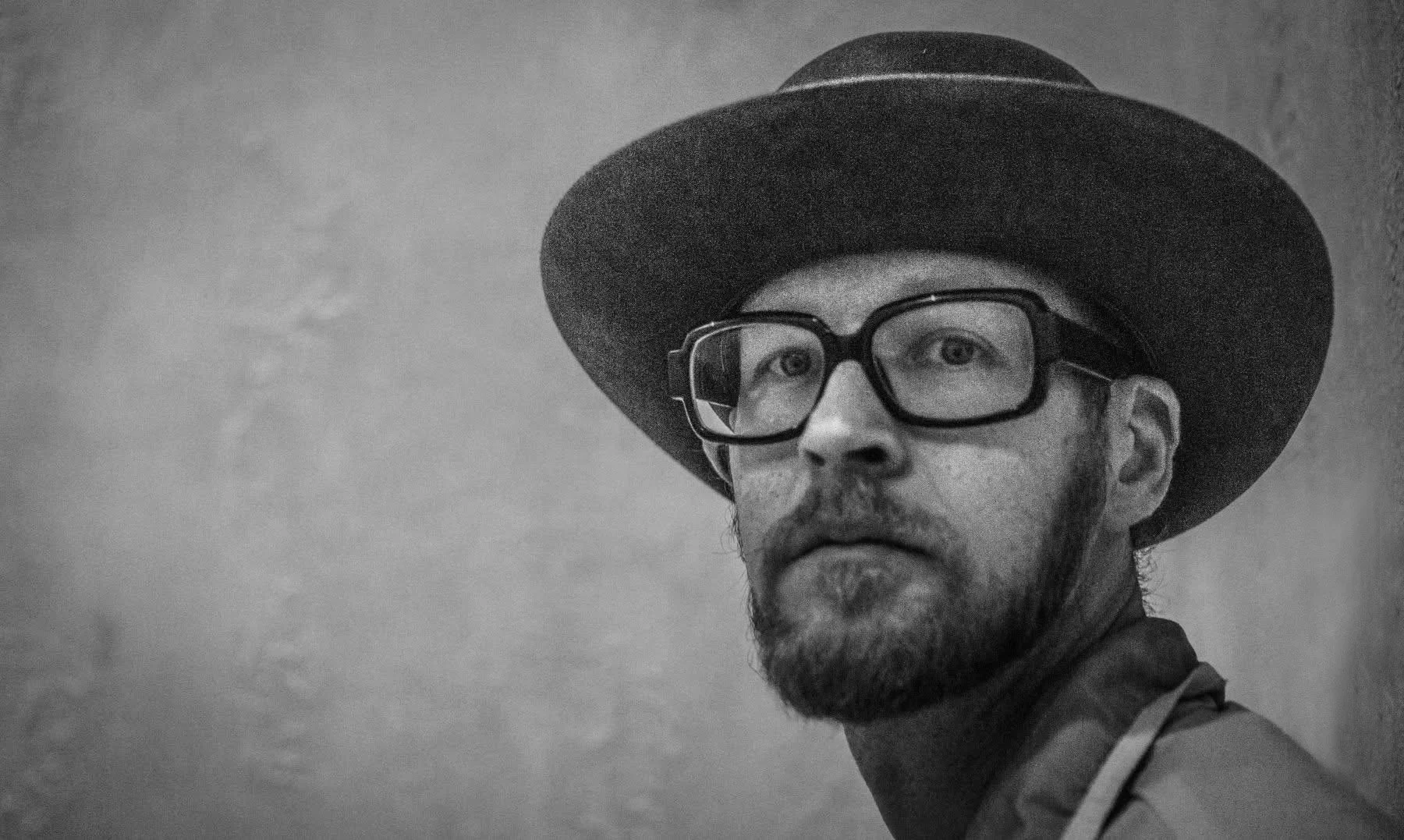 A black and white portrait of Kyle Van Osdol with a beard and glasses, wearing a wide-brimmed hat, looking to the side with an intense expression at Meow Wolf in Santa Fe, New Mexico.