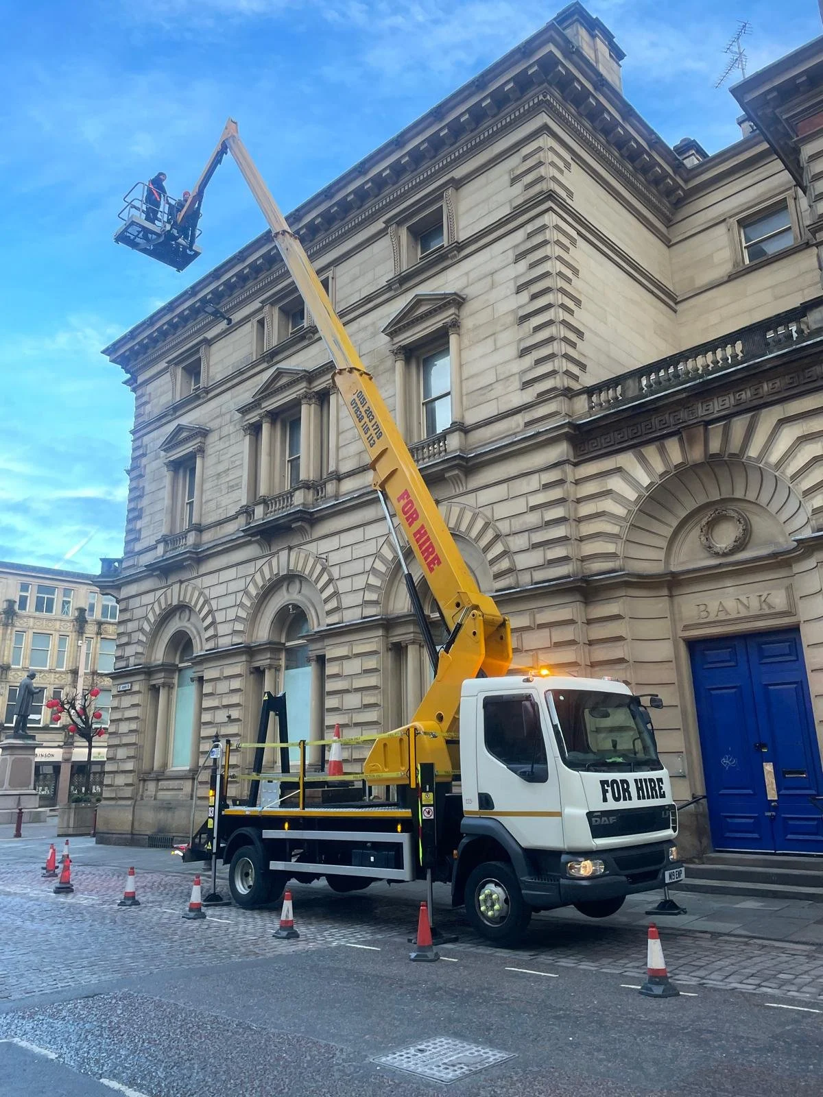 A person is using a hydraulic lift on a truck labeled 'FOR HIRE' to perform maintenance or inspection on a building's exterior, which has classical architectural details and a bank entrance. Orange cones outline a safety perimeter around the setup.