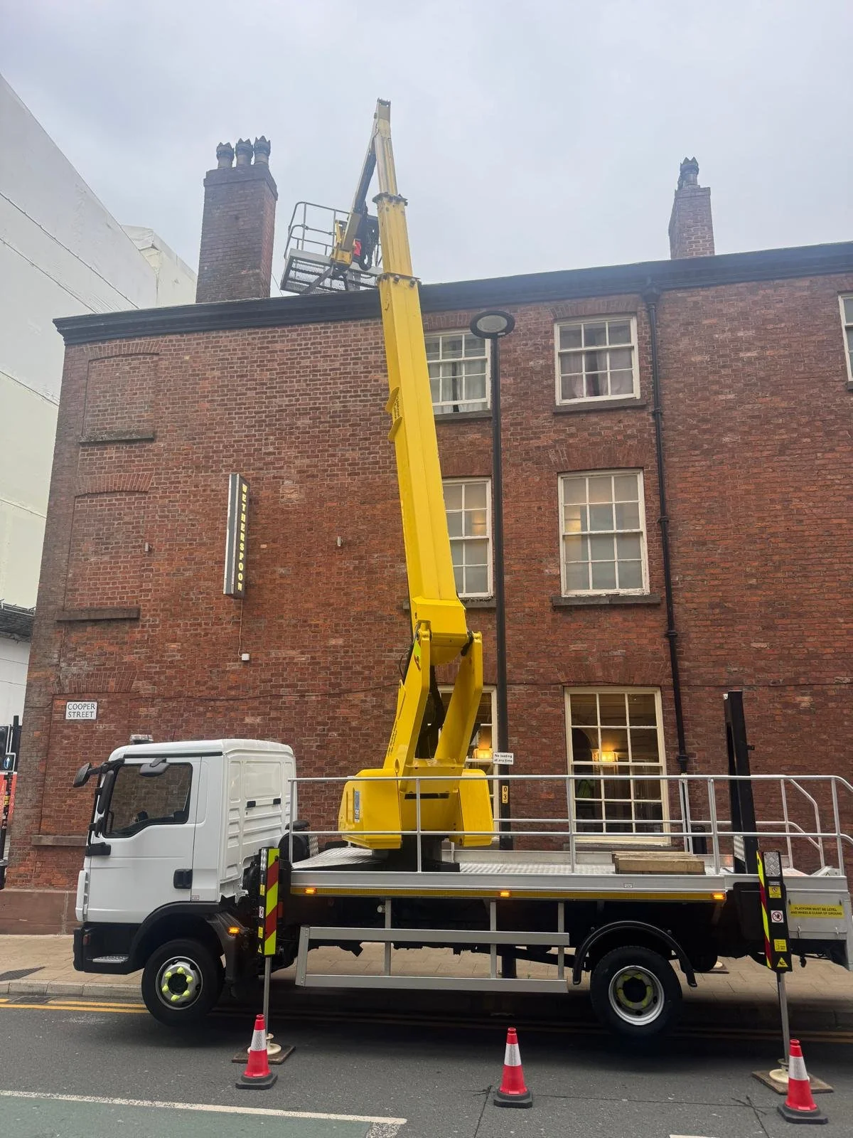 A white utility truck with a yellow aerial lift extends to reach the roof of a brick building. Orange traffic cones surround the truck, indicating park or work zone. The building has multiple windows and two prominent chimneys, with a street sign labeled 'Cooper Street' visible on the corner.