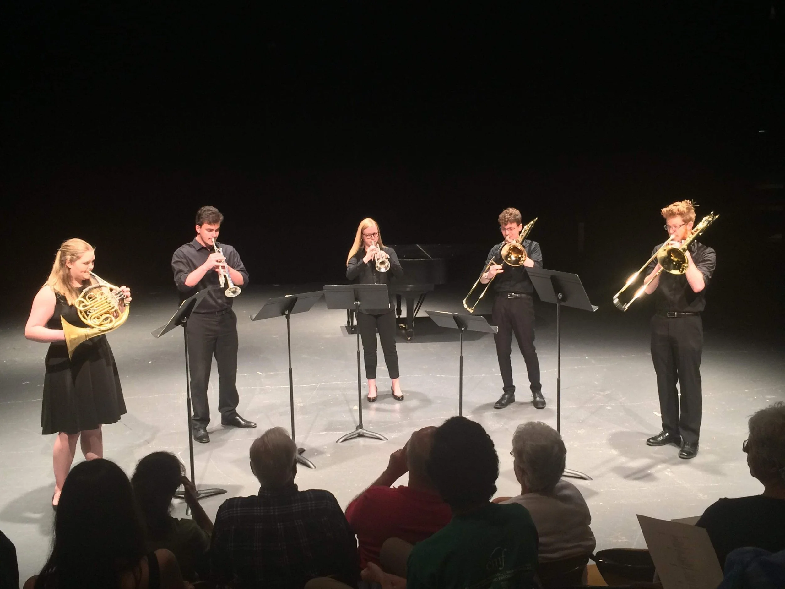 Five young musicians are performing on stage, playing various wind instruments including French horns, clarinets, and trombones, with music stands in front of them, in a darkened theater setting.