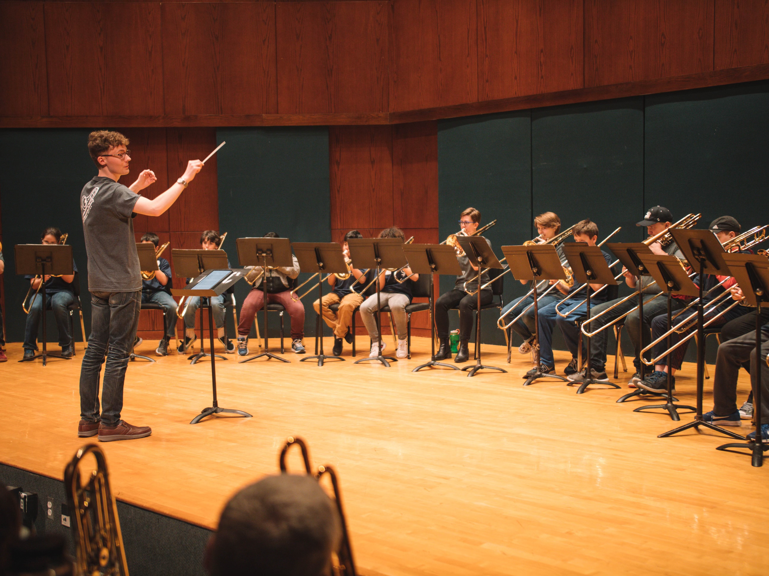 A conductor leading a band of young musicians on stage with brass and woodwind instruments, wooden paneling background.