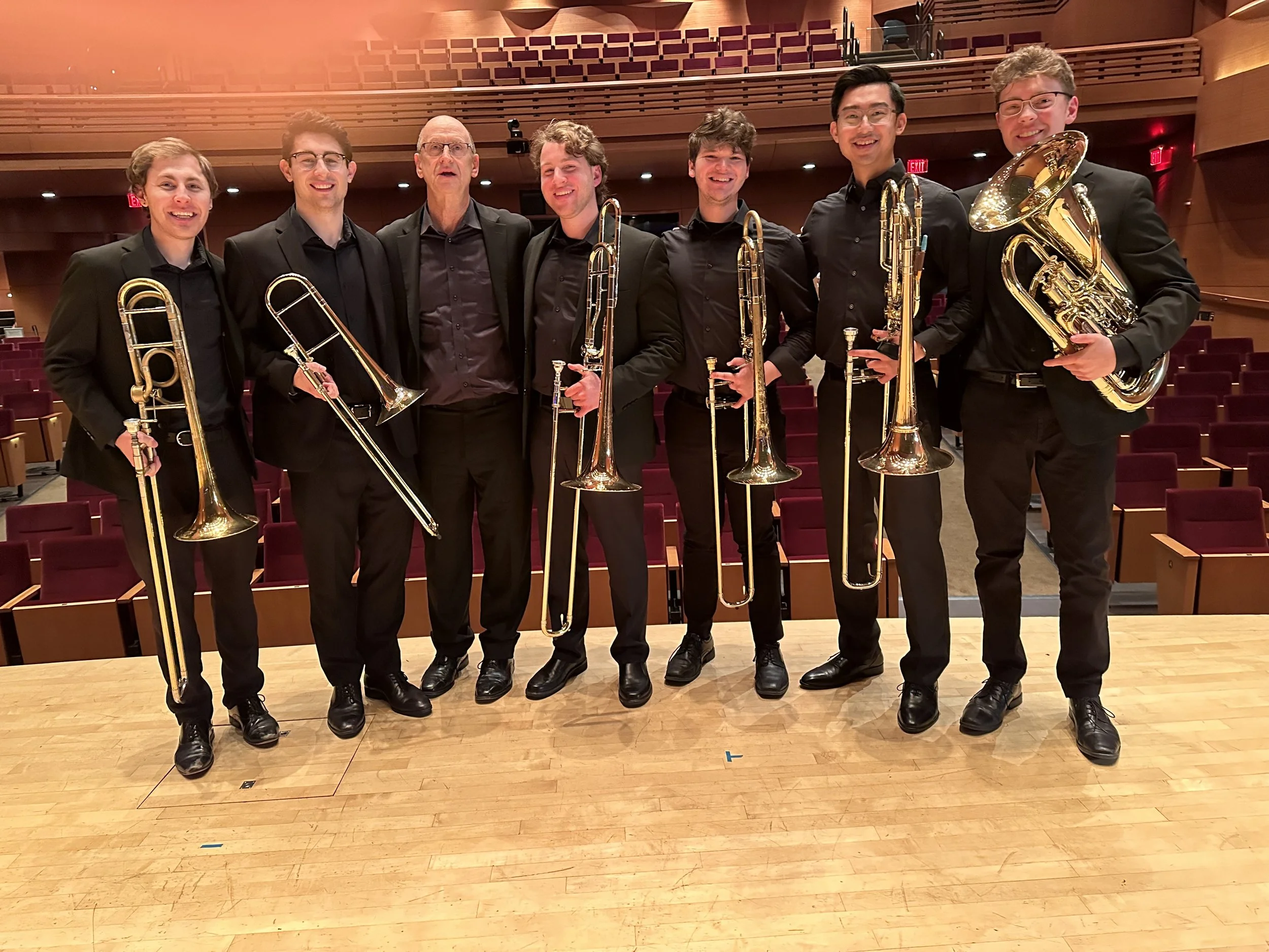 Group of six male musicians in black suits standing in a concert hall, holding trombones and euphoniums, smiling at the camera.