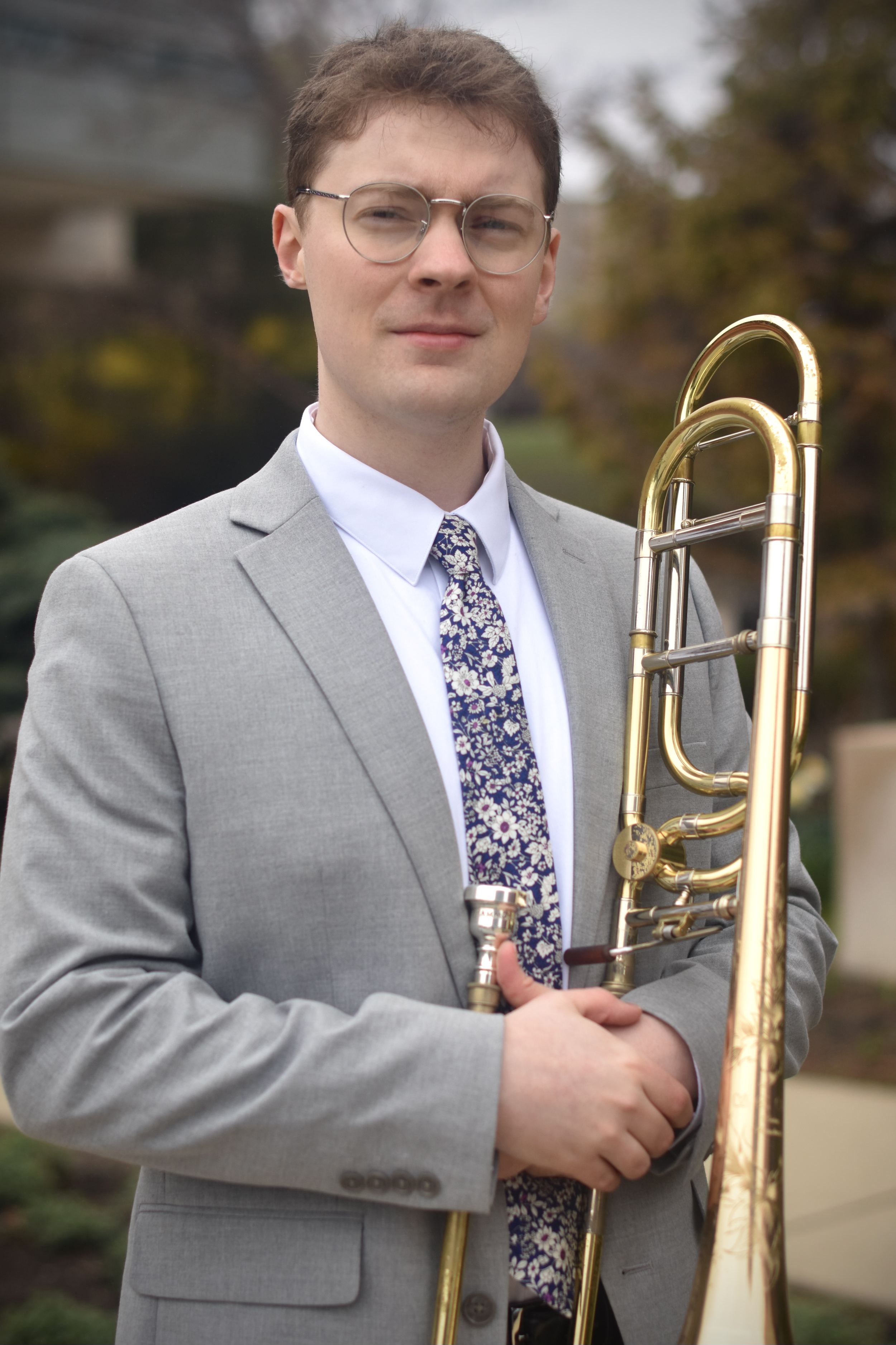 A man wearing a gray suit, glasses, and a floral tie holds a trombone outdoors during daytime.