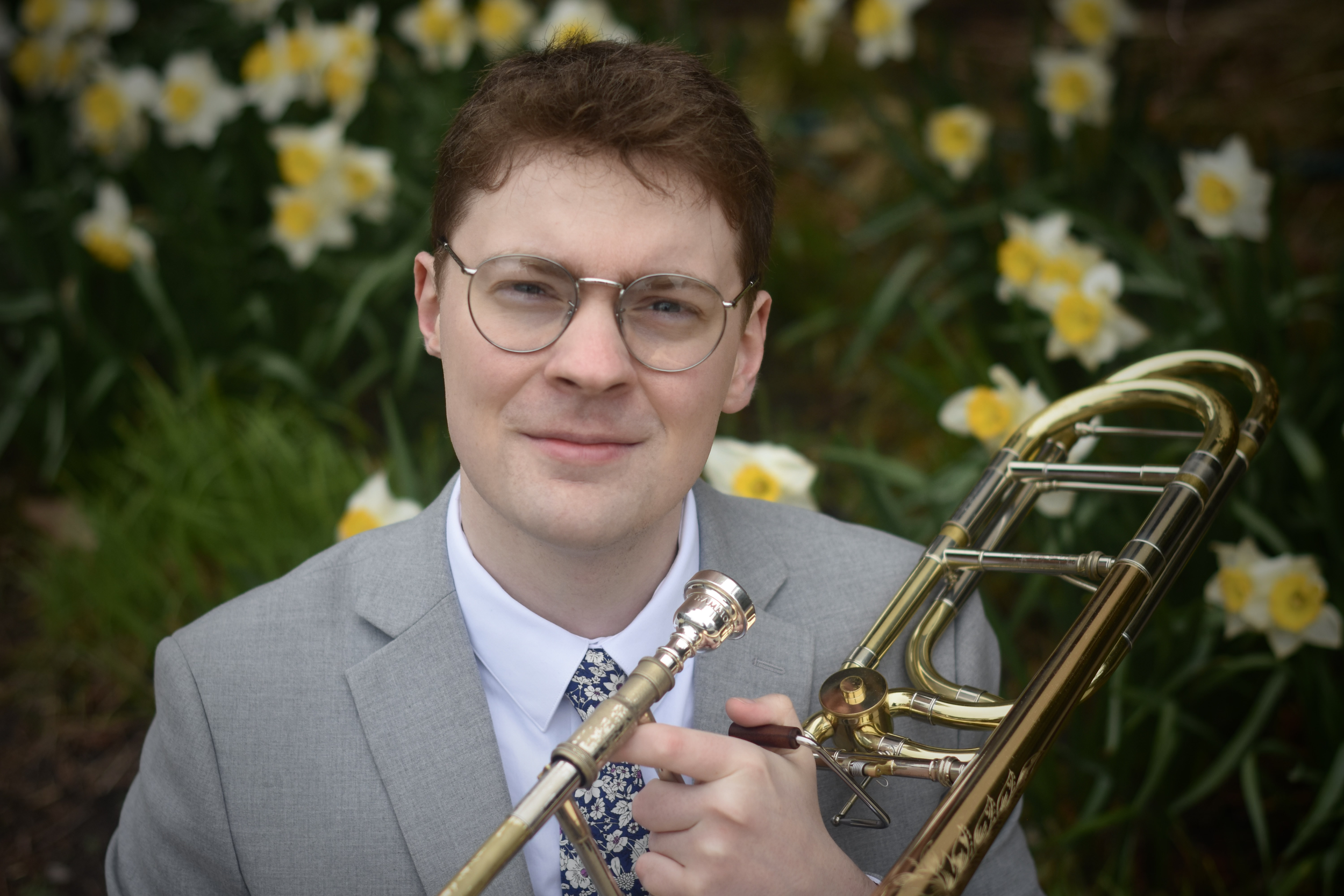 A young man with brown hair, glasses, and wearing a gray suit and patterned tie playing a saxophone outdoors, surrounded by white and yellow flowers.