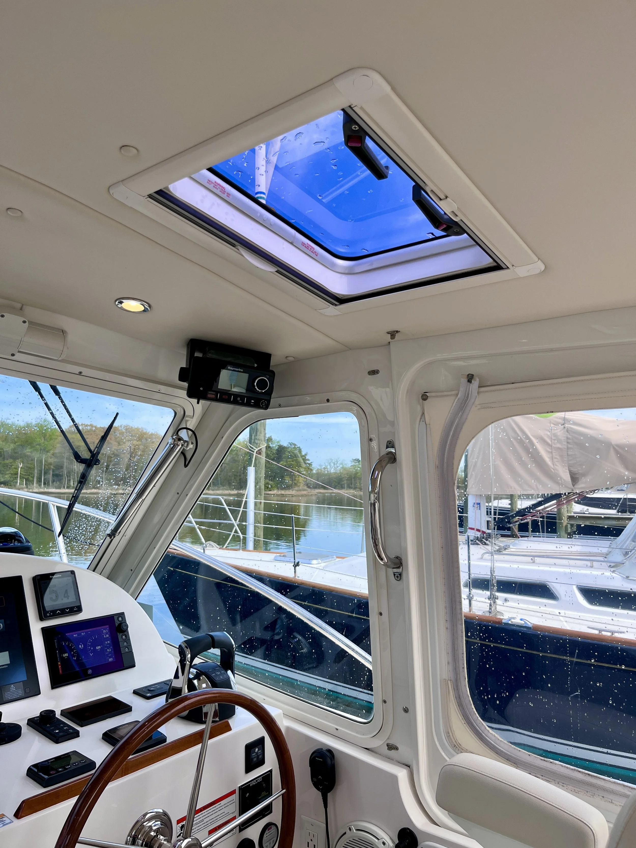 The image shows the interior of a boat's cockpit area with various navigation instruments and a large window with water droplets on it. There is a skylight on the ceiling showing a blue sky with some rain drops, and outside the window, other boats ar