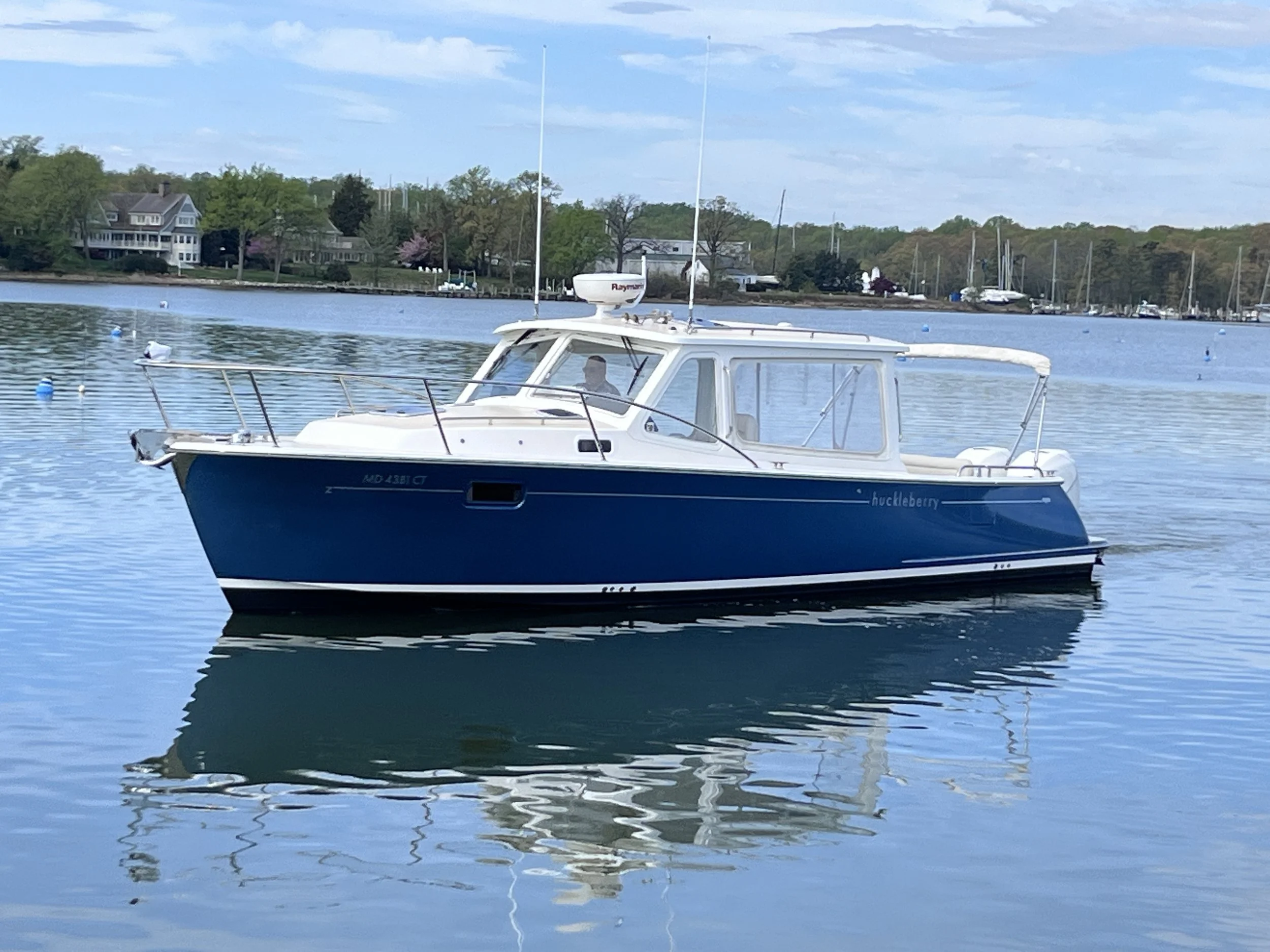 A blue and white boat with the name 'huckleberry' on its side floating on a calm body of water, with houses and trees in the background on a partly cloudy day.
