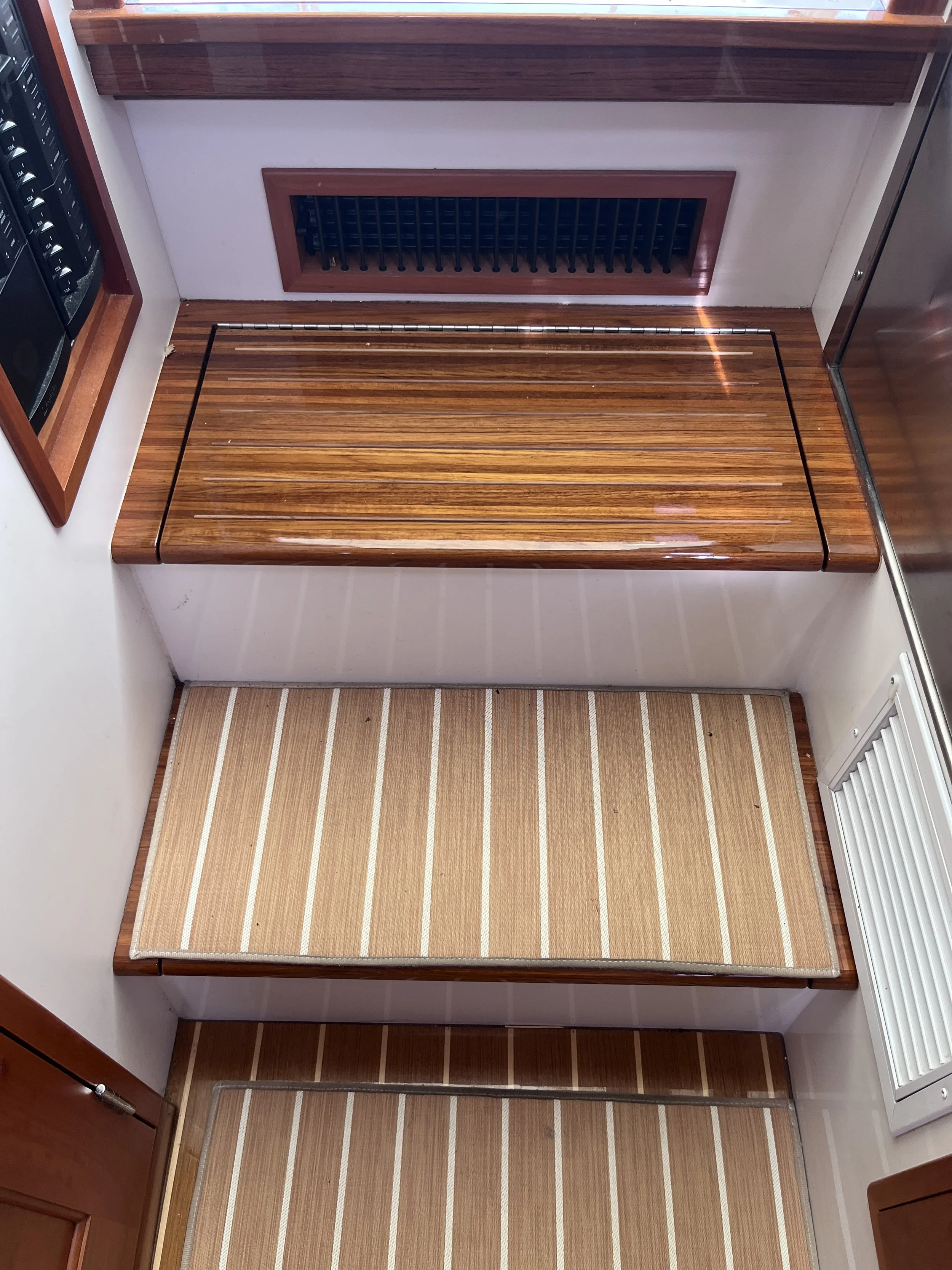 Interior view of a wooden boat with a striped deck and a wooden bench seat, a vent on the wall, and a window with a wooden frame.