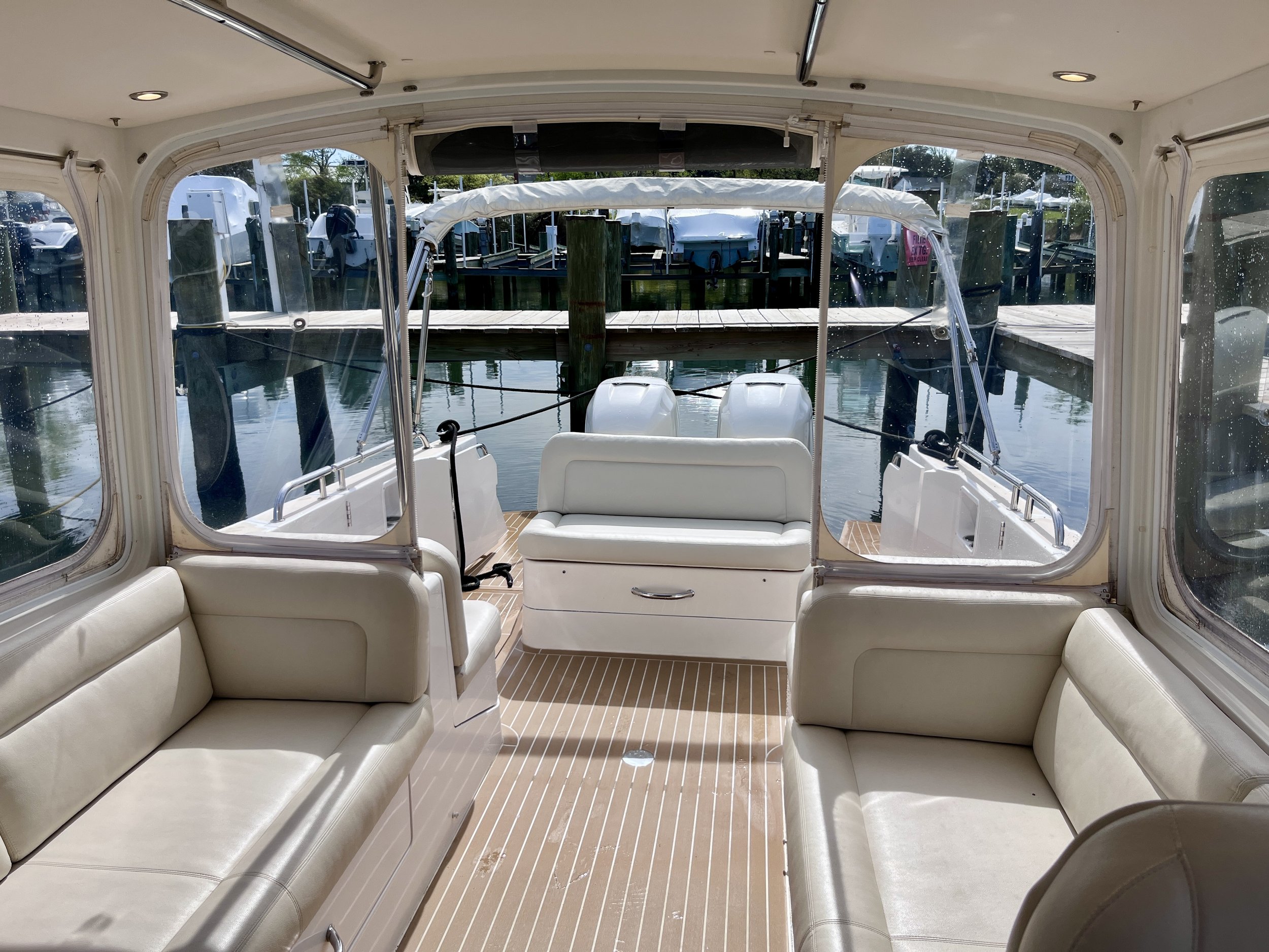 Interior view of a boat with cream-colored seating, viewed from the back looking forward, with a view of the dock and other boats outside through clear windows.