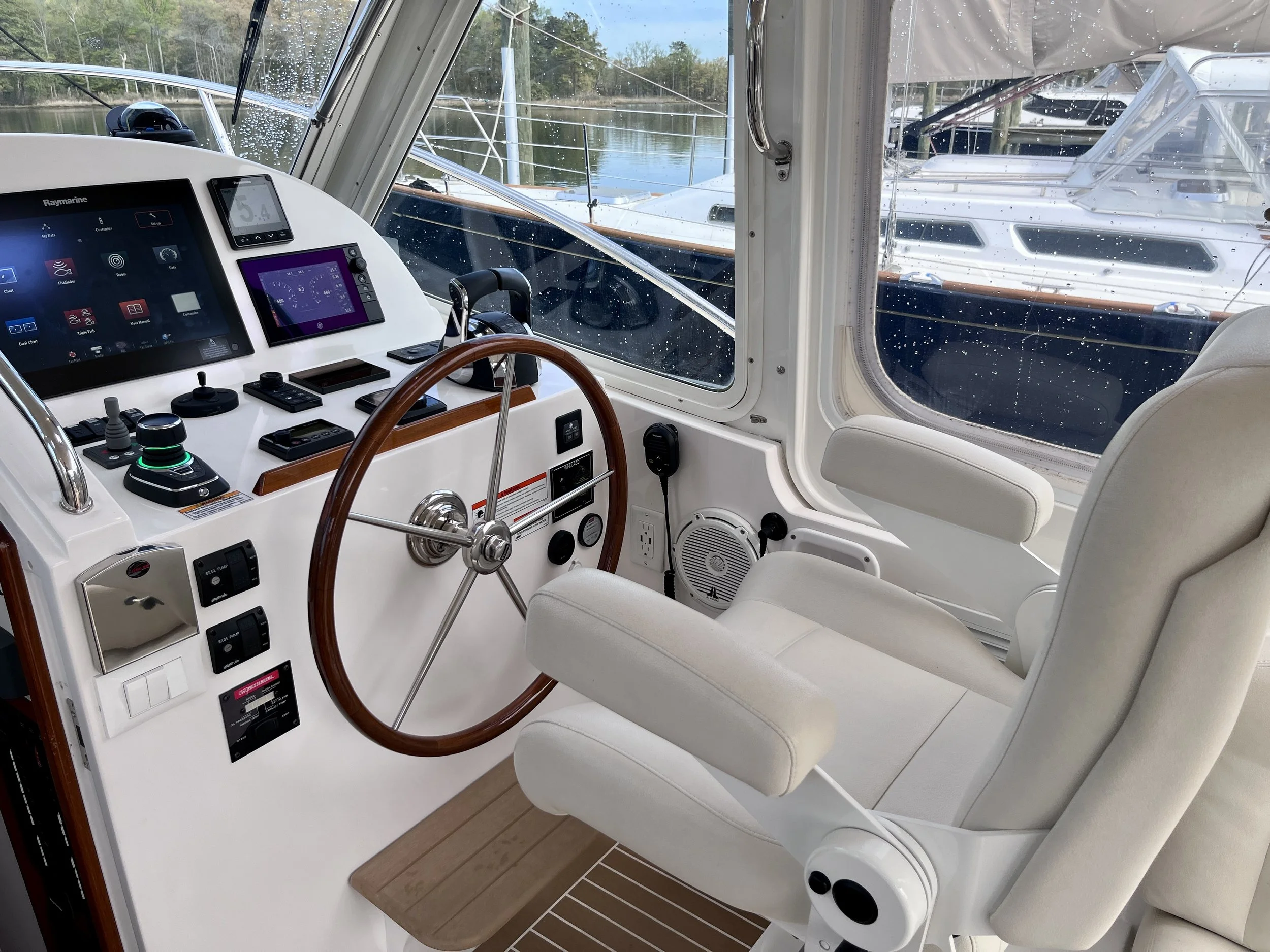The interior of a yacht's steering area with various navigational and control instruments, a wooden steering wheel, and two white cushioned chairs. Outside, through rain-covered windows, other boats are docked at a marina, with water and trees visibl