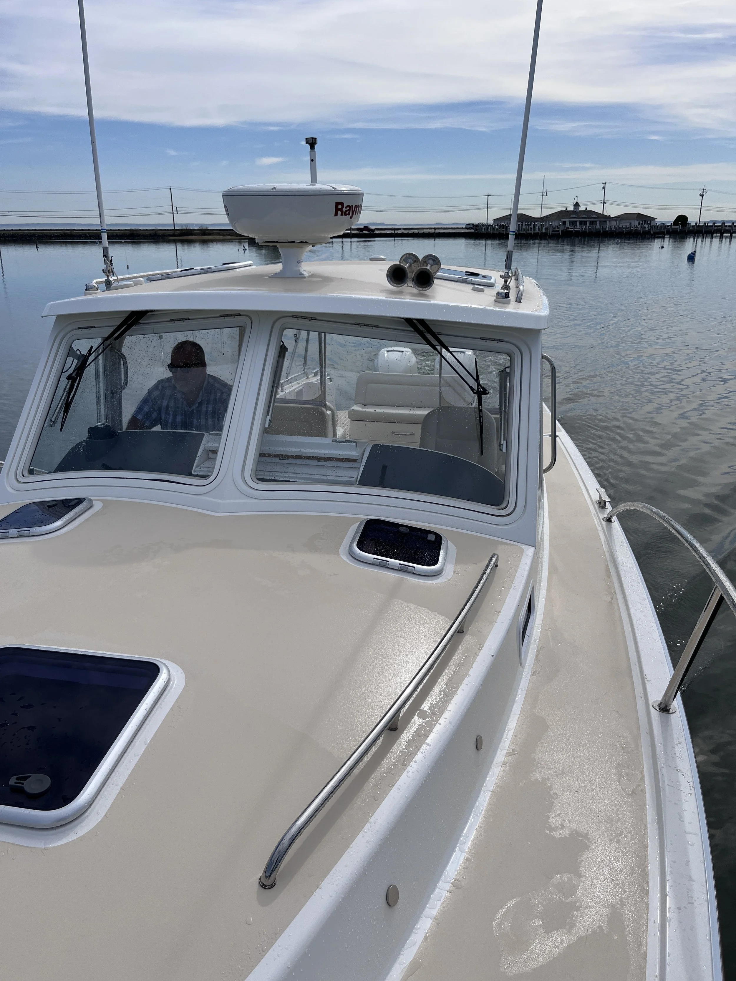 View of a boat on calm water with a man inside, looking at a device. The boat has a radar or navigation equipment on the roof and is surrounded by water with a dock and buildings in the background under a partly cloudy sky.