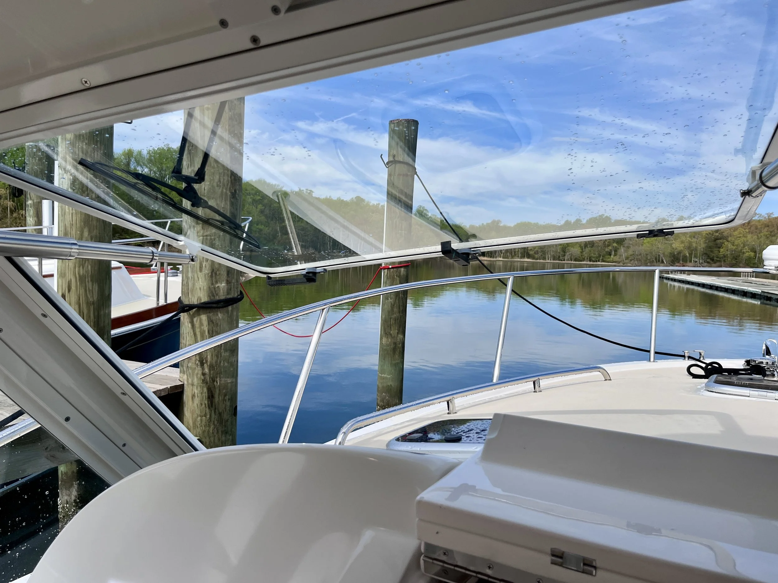 View from inside a boat with a clear glass roof, looking out over a wooden dock and water with trees in the background on a partly cloudy day.