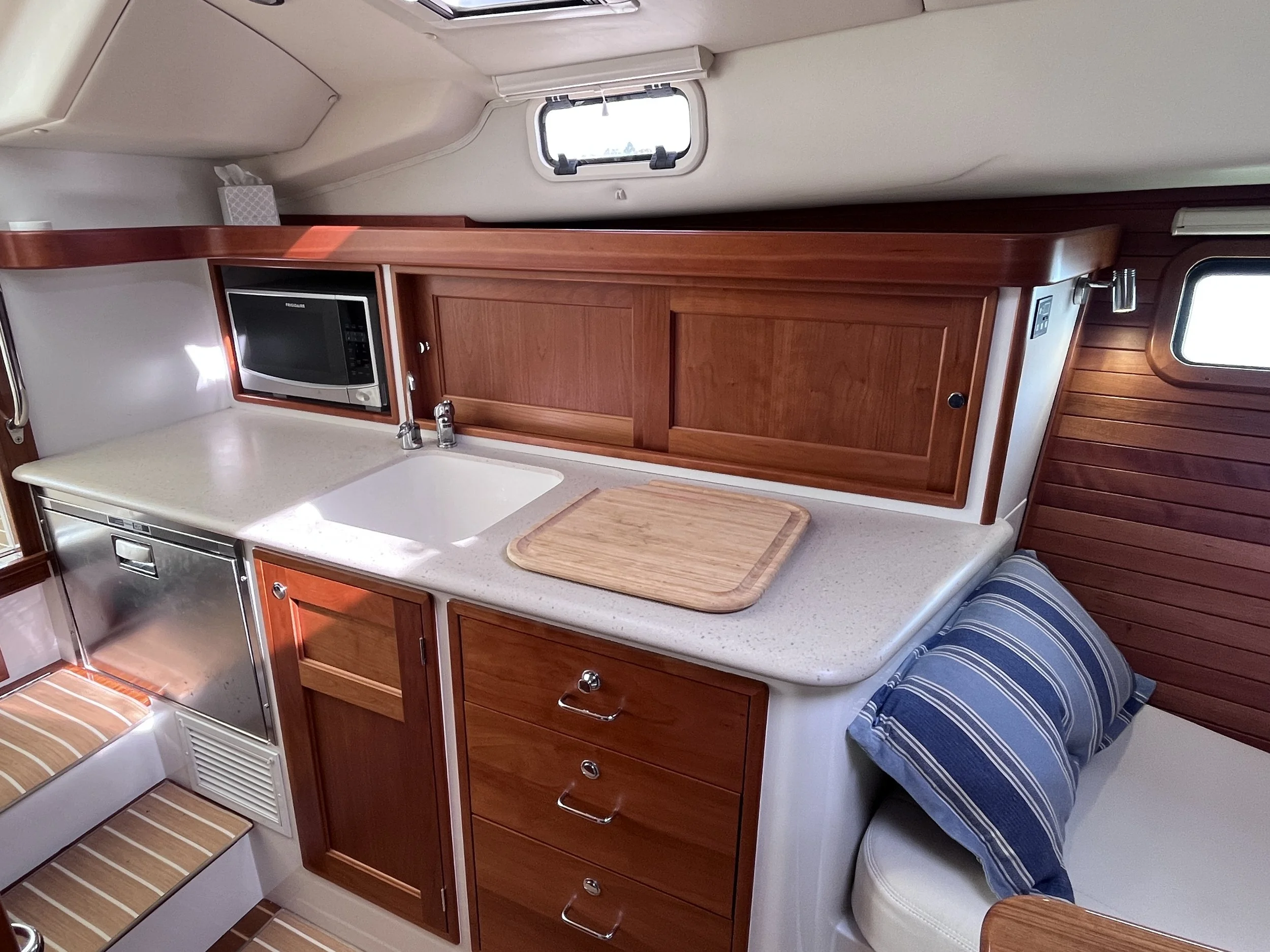 Interior of a boat or yacht kitchen with wooden cabinetry, a microwave, a white countertop, a sink, and a small window above.