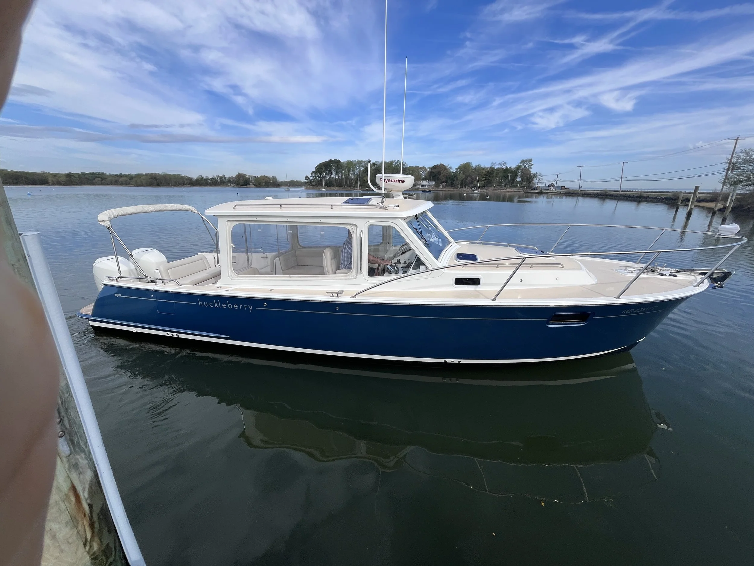 A white and blue motorboat named 'huckleberry' floating on calm water with trees, boats, and power lines in the background and a partly cloudy sky overhead.
